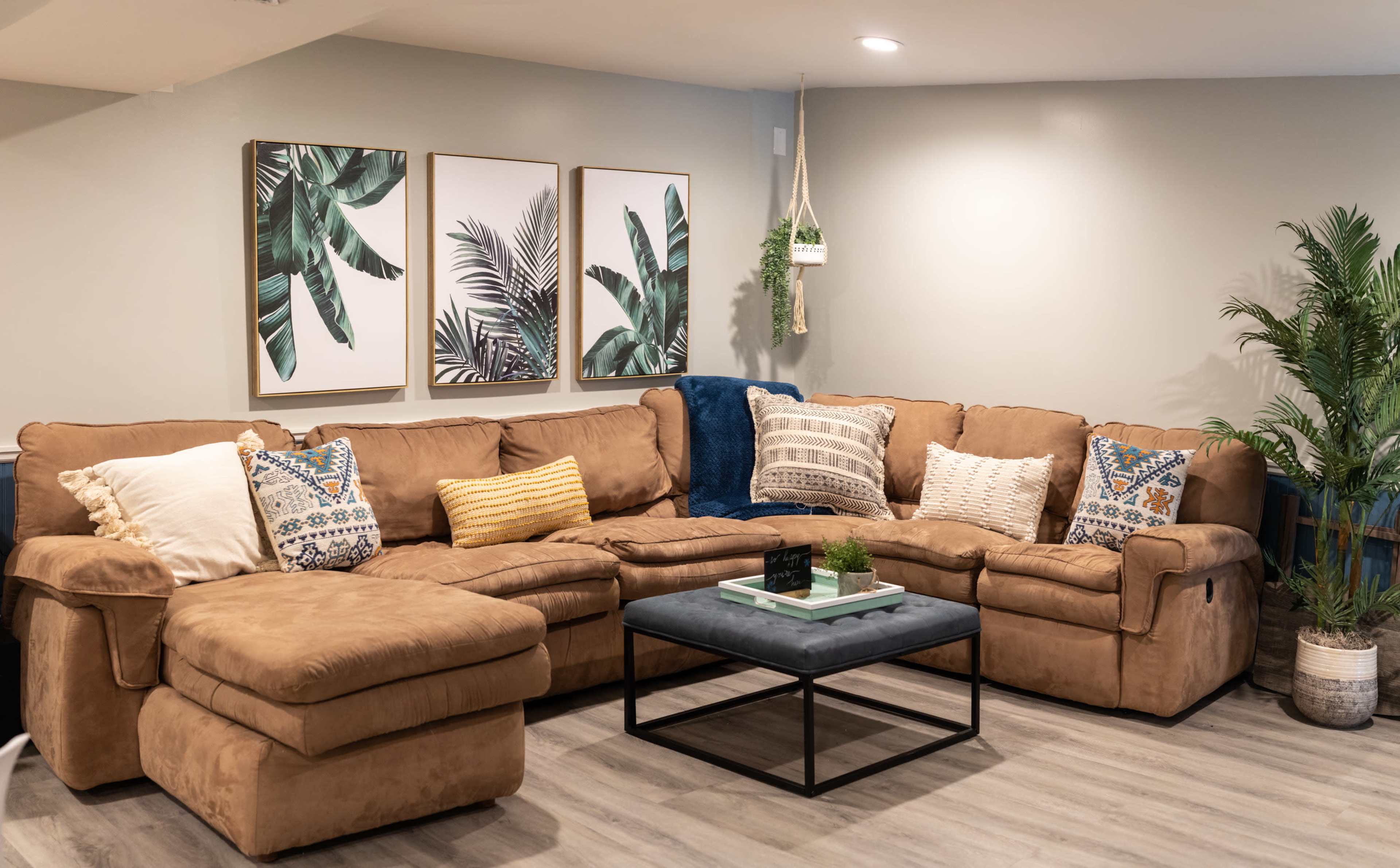 A cozy living room features a large brown sectional sofa arranged around a low coffee table, complemented by wall art depicting tropical plants.