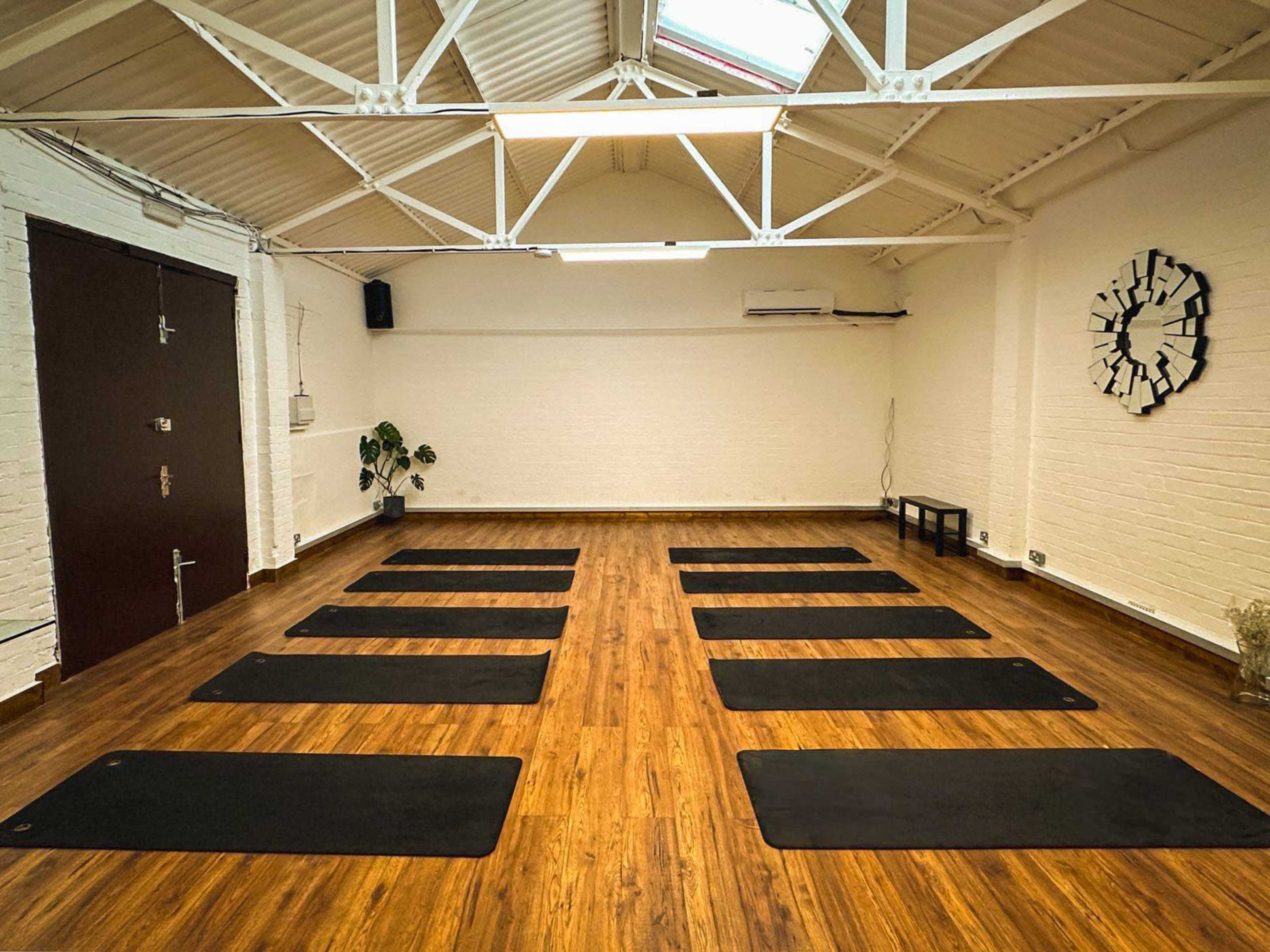The image shows a spacious yoga studio with wooden flooring, featuring several yoga mats arranged neatly in rows, and minimalistic decor on the walls.
