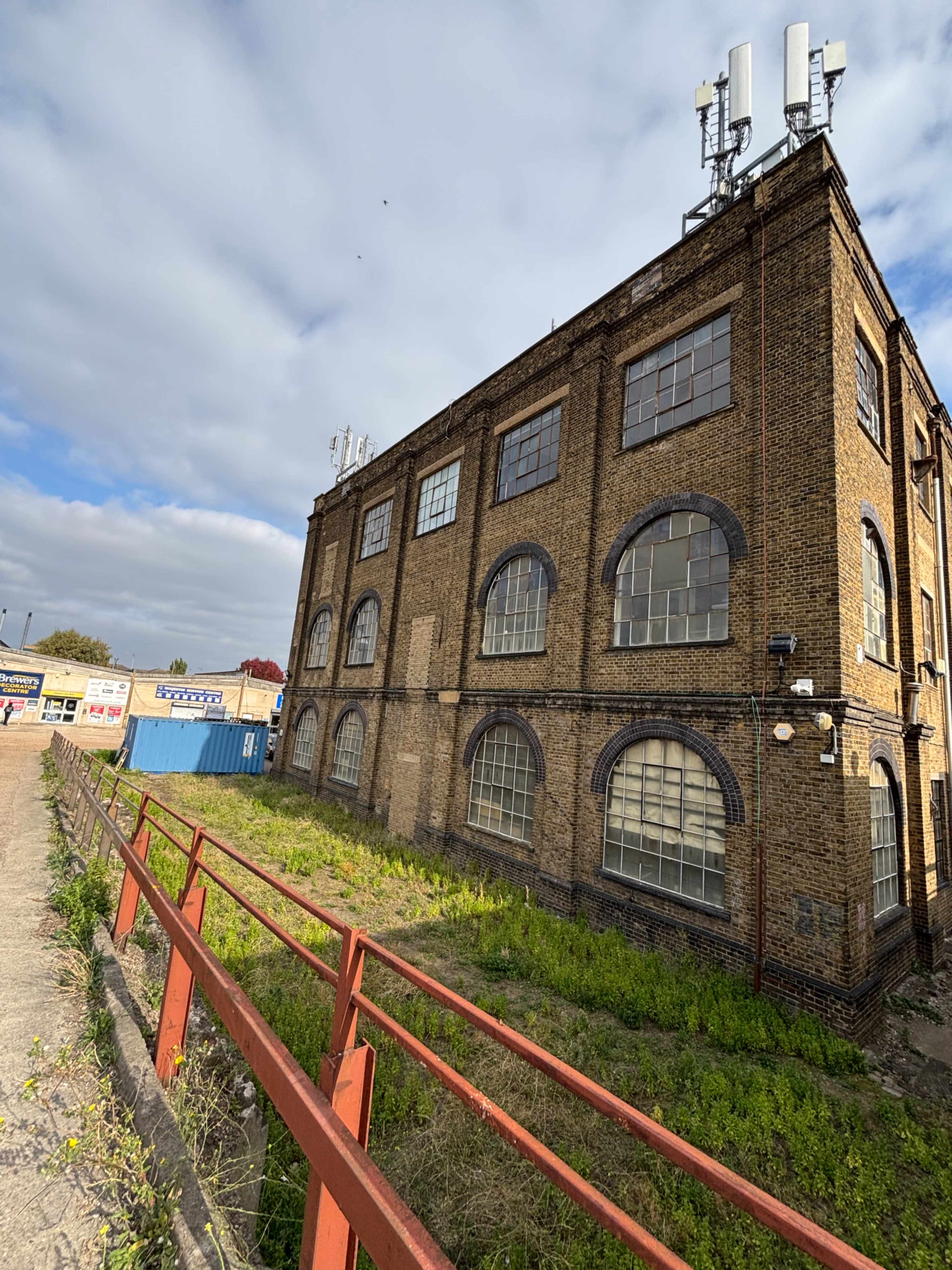 The image shows a brick industrial building with arched windows and several antennas on the roof, situated beside a grassy area and a pathway.