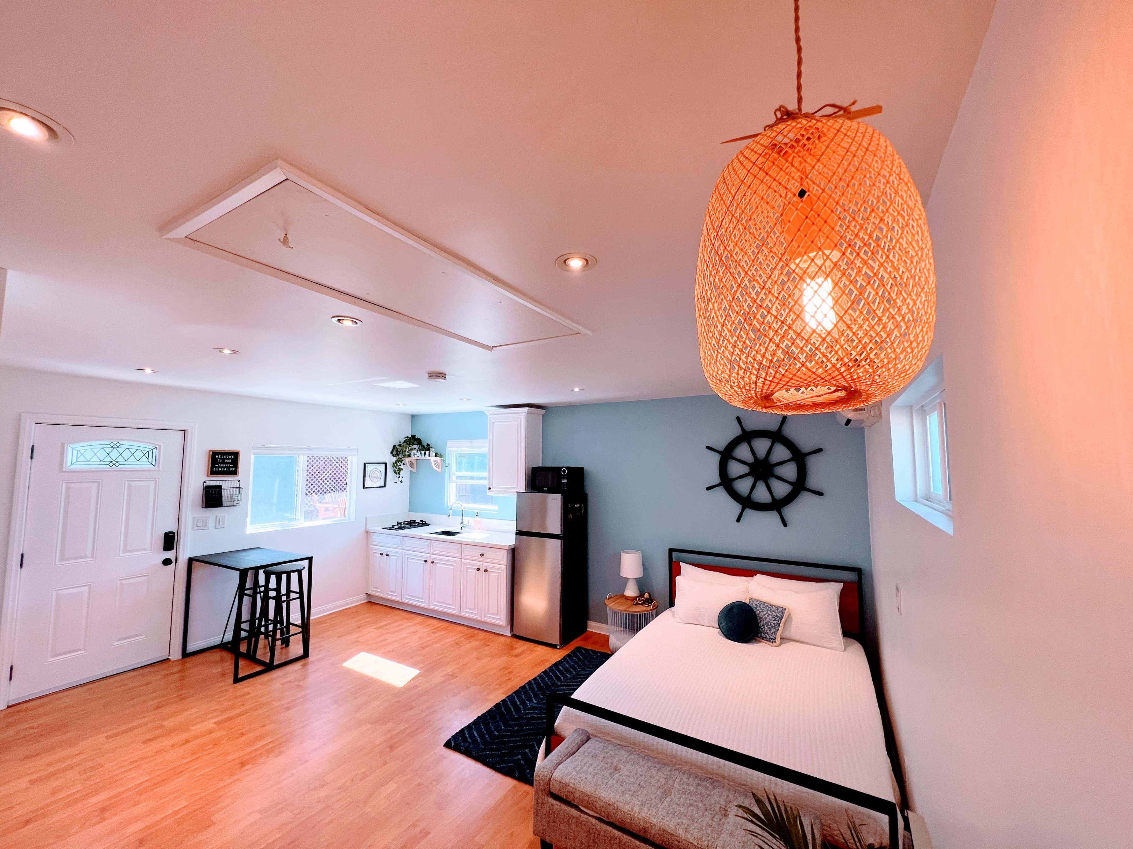 The image showcases a bright, open studio apartment featuring a bed, small dining area, kitchenette, and nautical-themed decor.