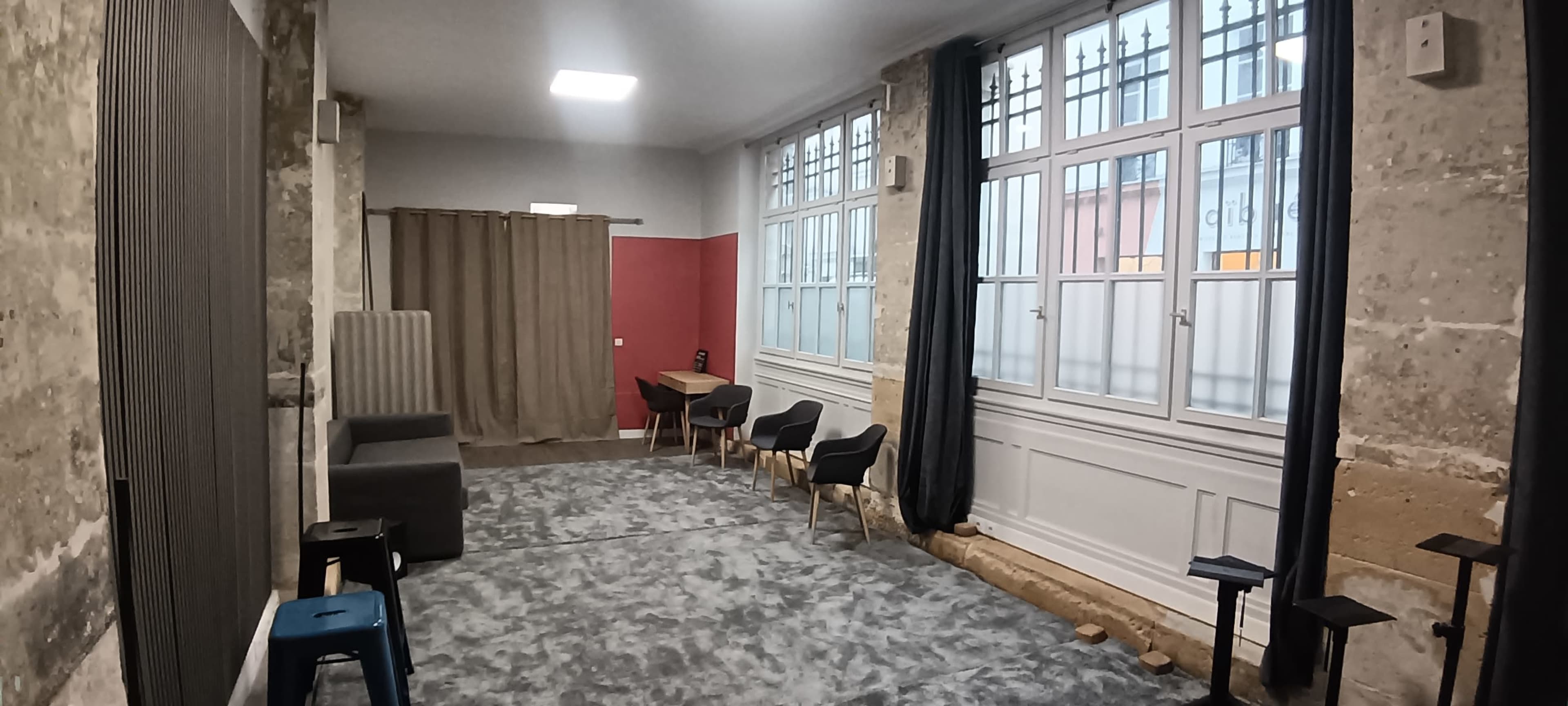 The image shows an interior space with stone walls, large windows, a gray carpet, and furniture including chairs and a table.