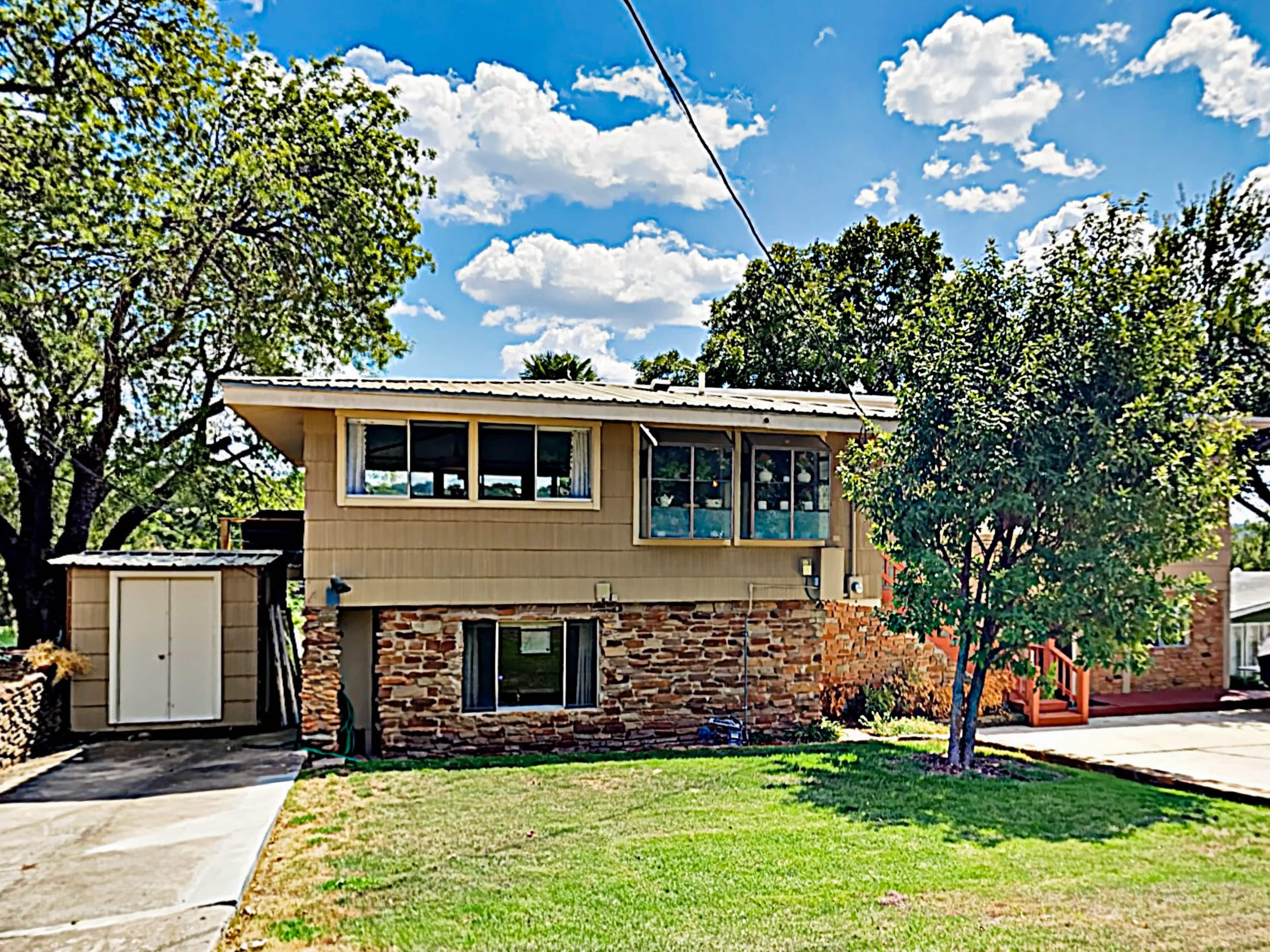 The image shows a two-story house with a combination of stone and wood siding, surrounded by a green lawn and trees under a sunny sky.