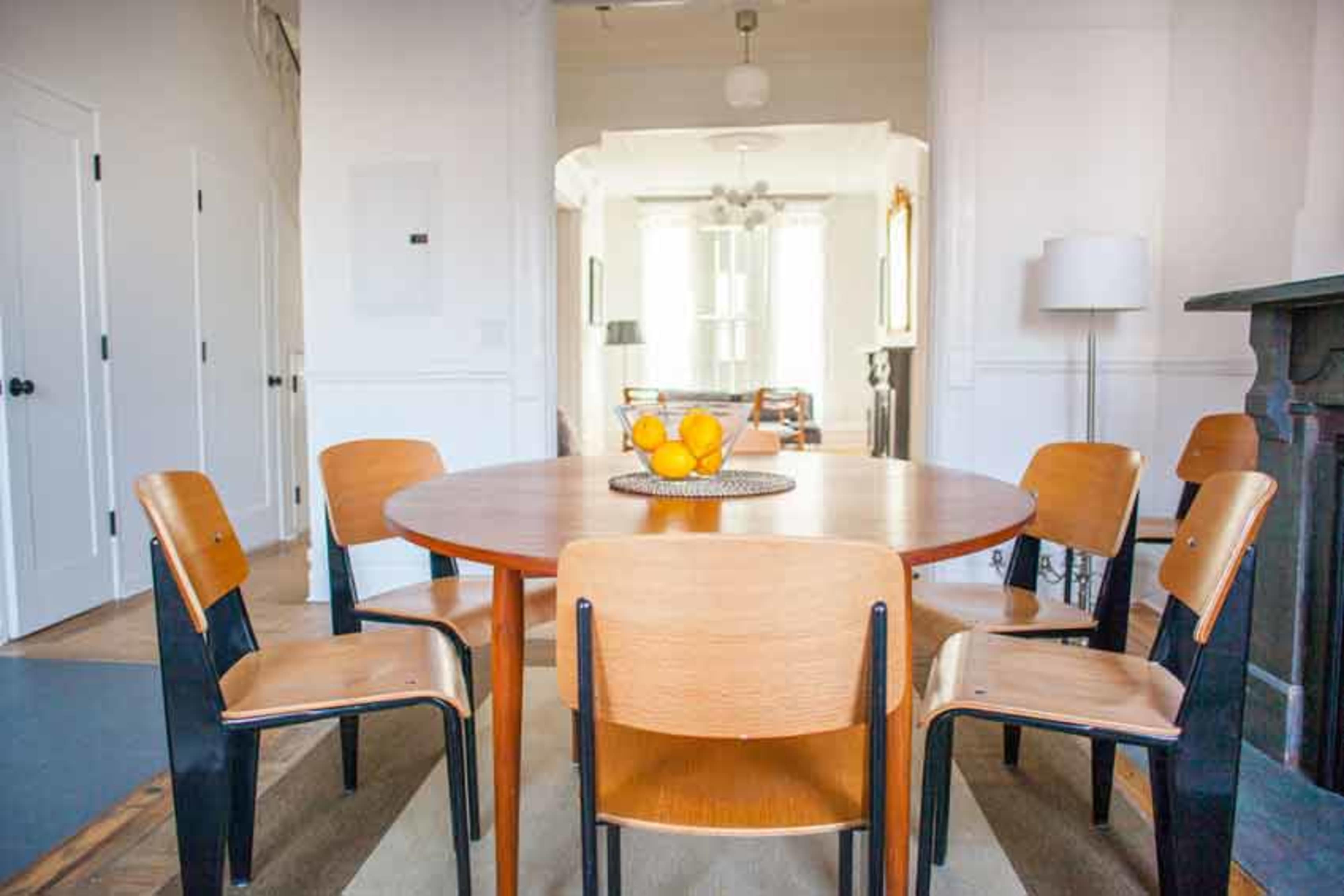 A round wooden dining table with six chairs surrounds a bowl of oranges, set in a well-lit room with white walls and doorways leading to other areas.