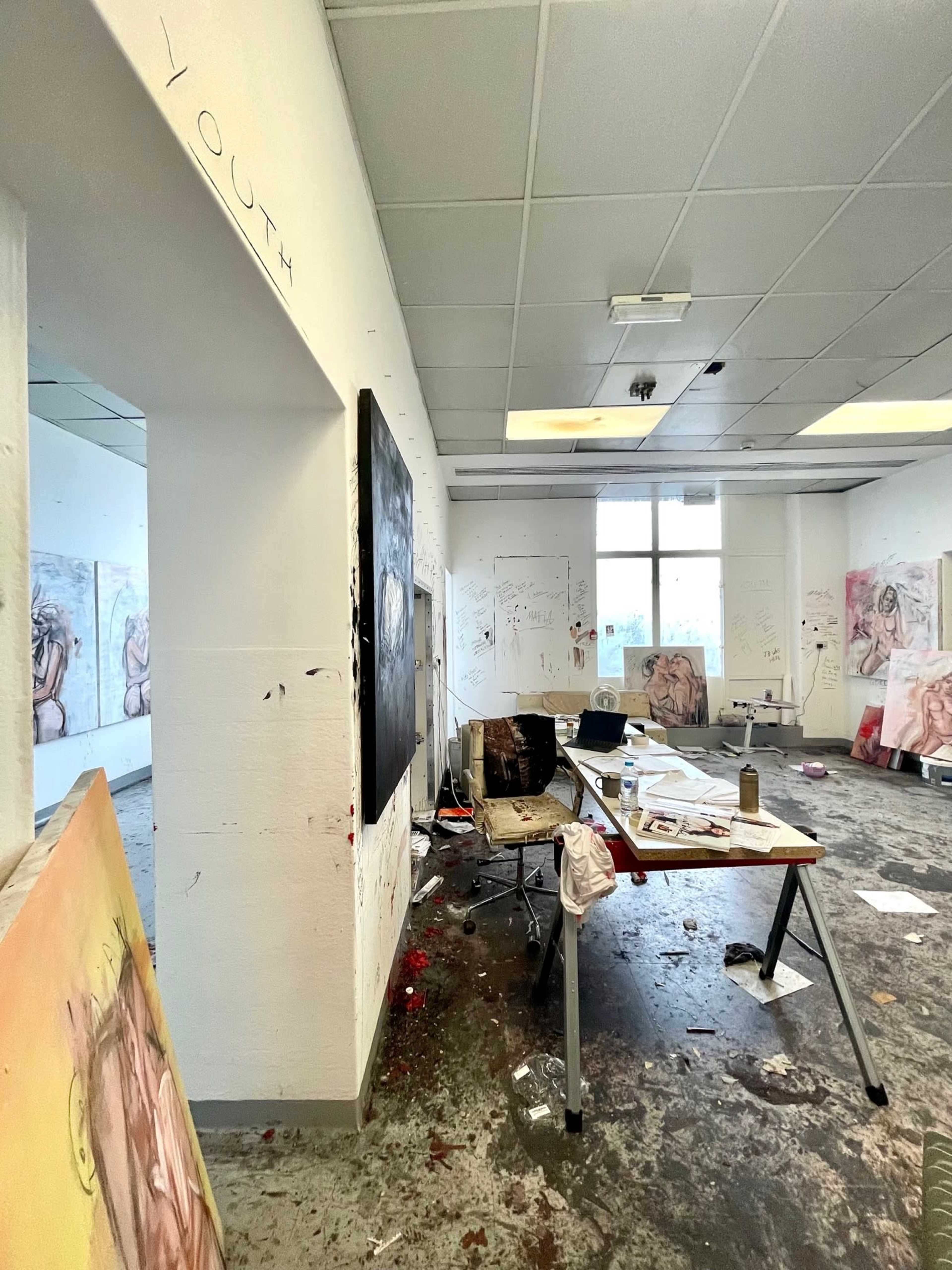 A cluttered art studio with scattered artworks, paint stains on the floor, and a partially furnished workspace.