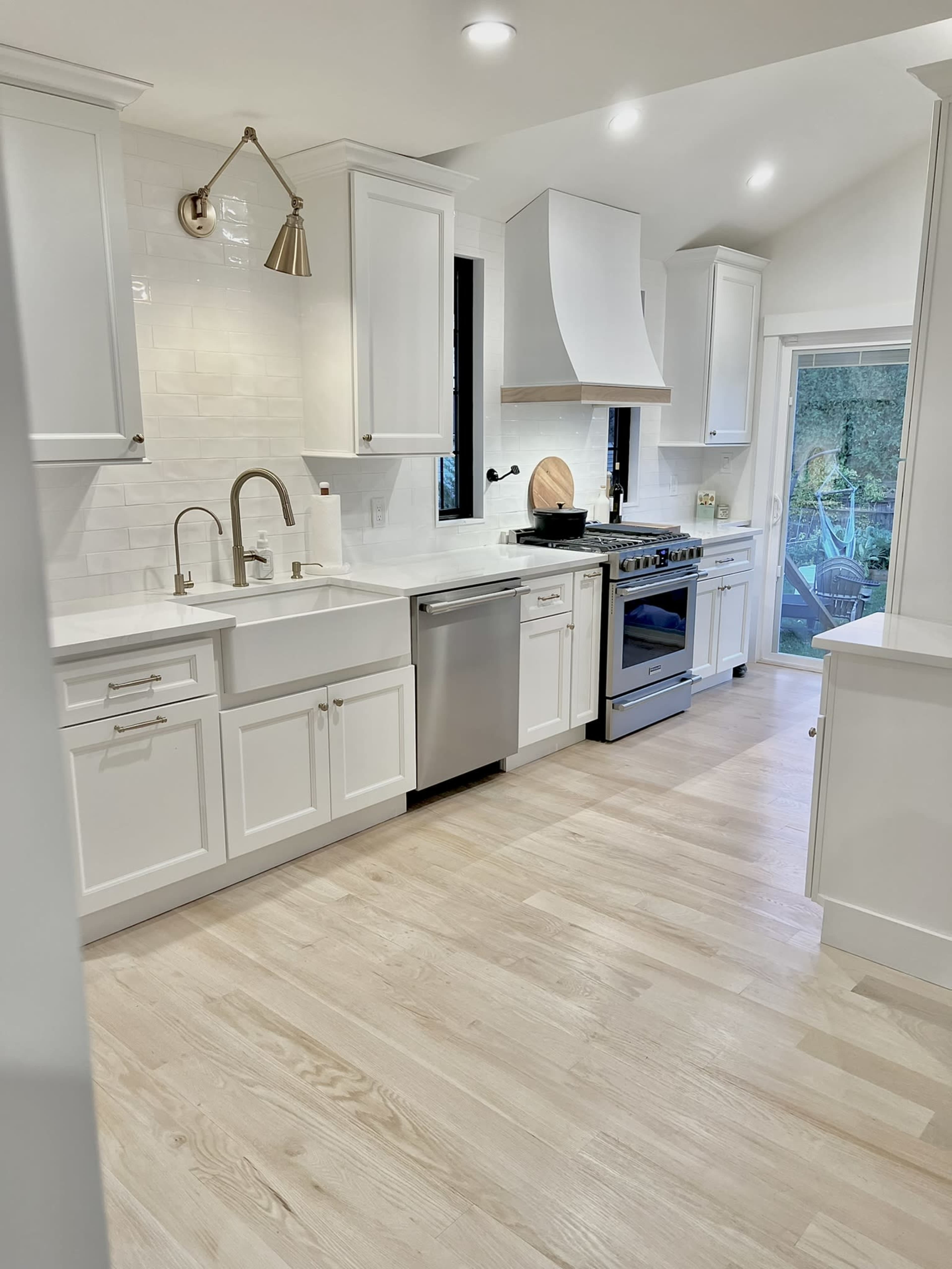 A modern kitchen with white cabinets, a stainless steel dishwasher, and a silver stove, with a view of the outdoors through a glass door.