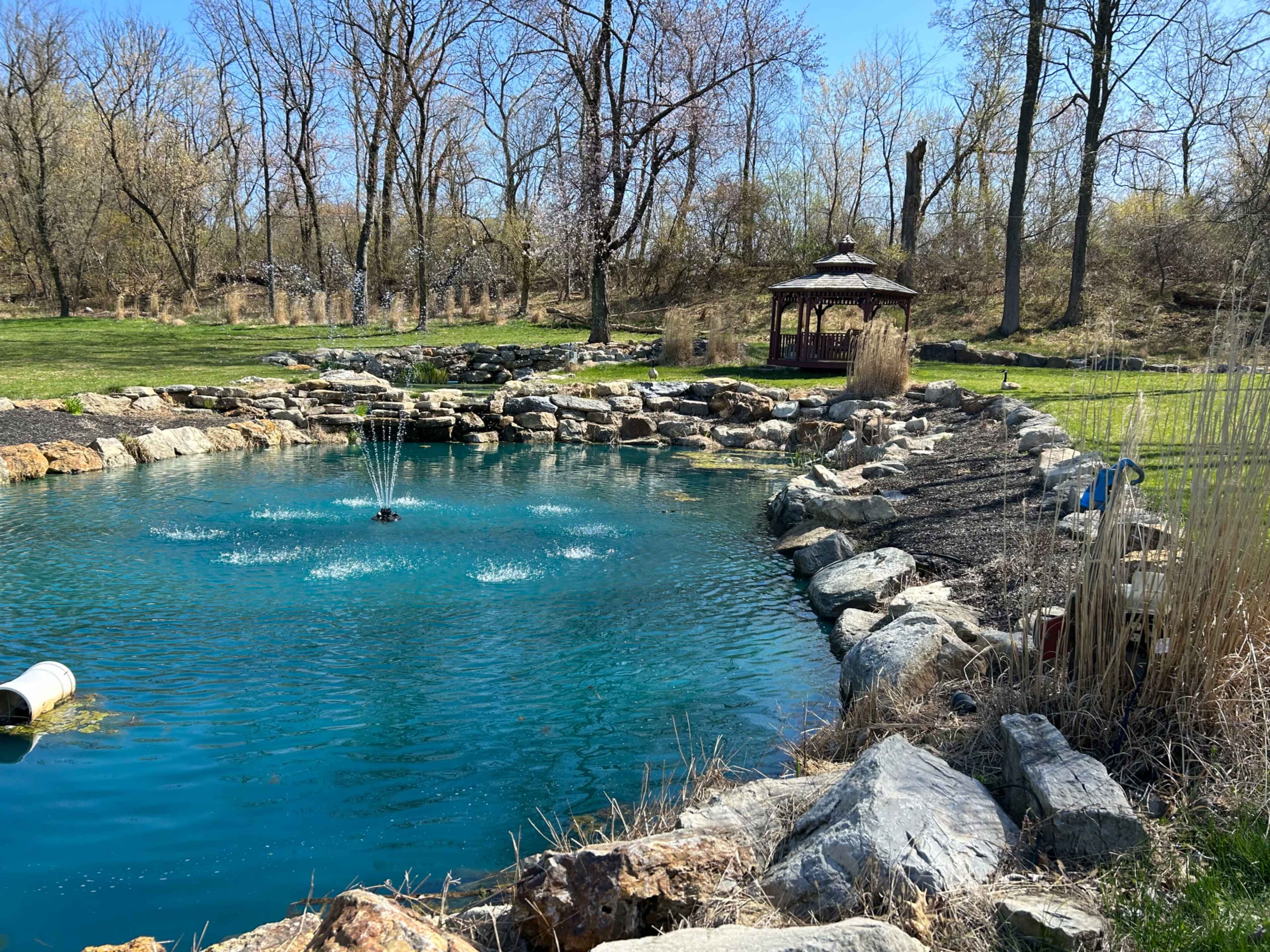 The image shows a pond with a fountain surrounded by rocks and a gazebo with trees in the background.