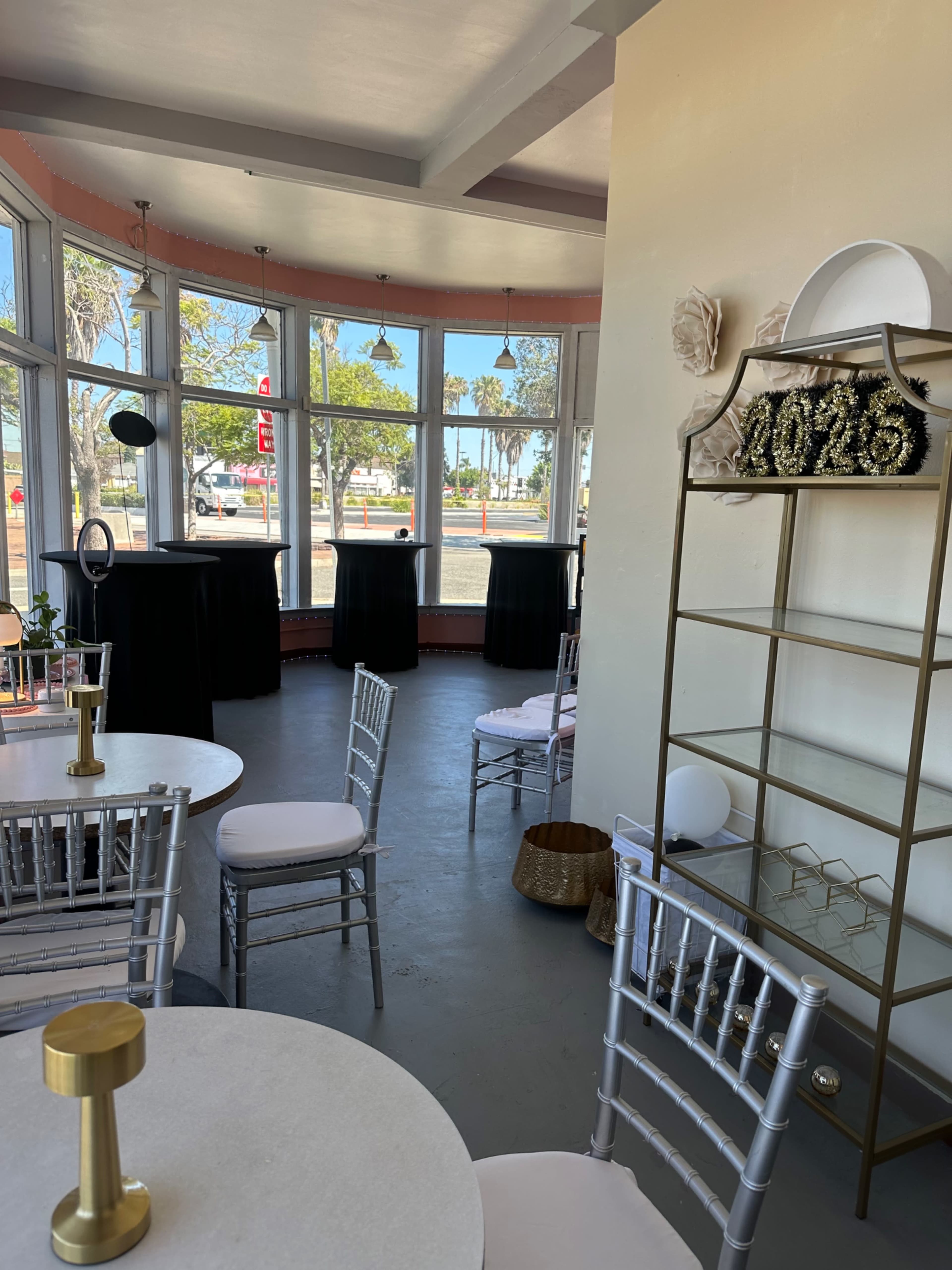 The image shows a small event space featuring tables with white chairs, large windows with natural light, and a decorative shelf with items displayed.