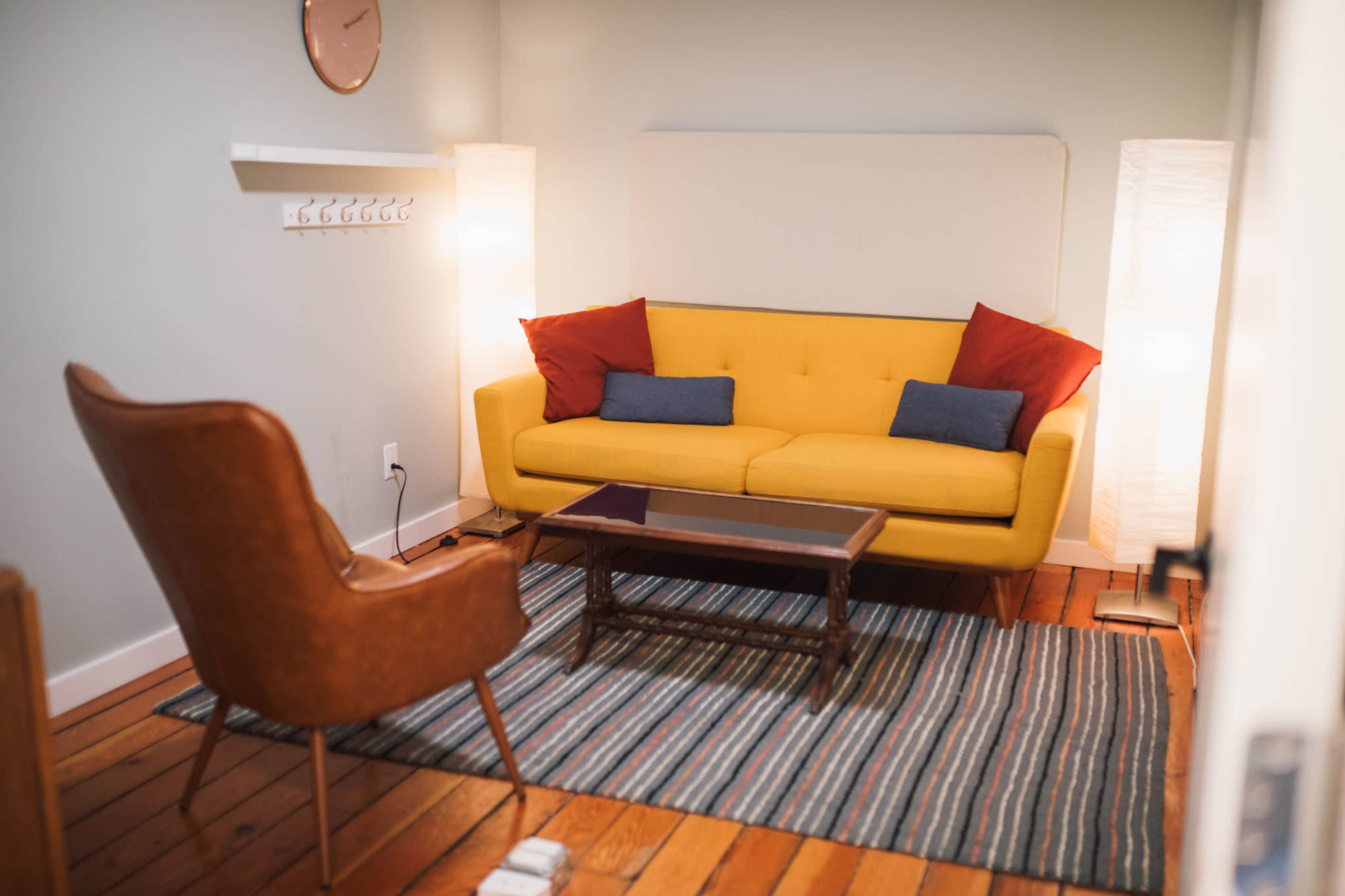 The image shows a cozy living room with a yellow sofa, two floor lamps, a wooden coffee table, a patterned rug, and a brown accent chair.