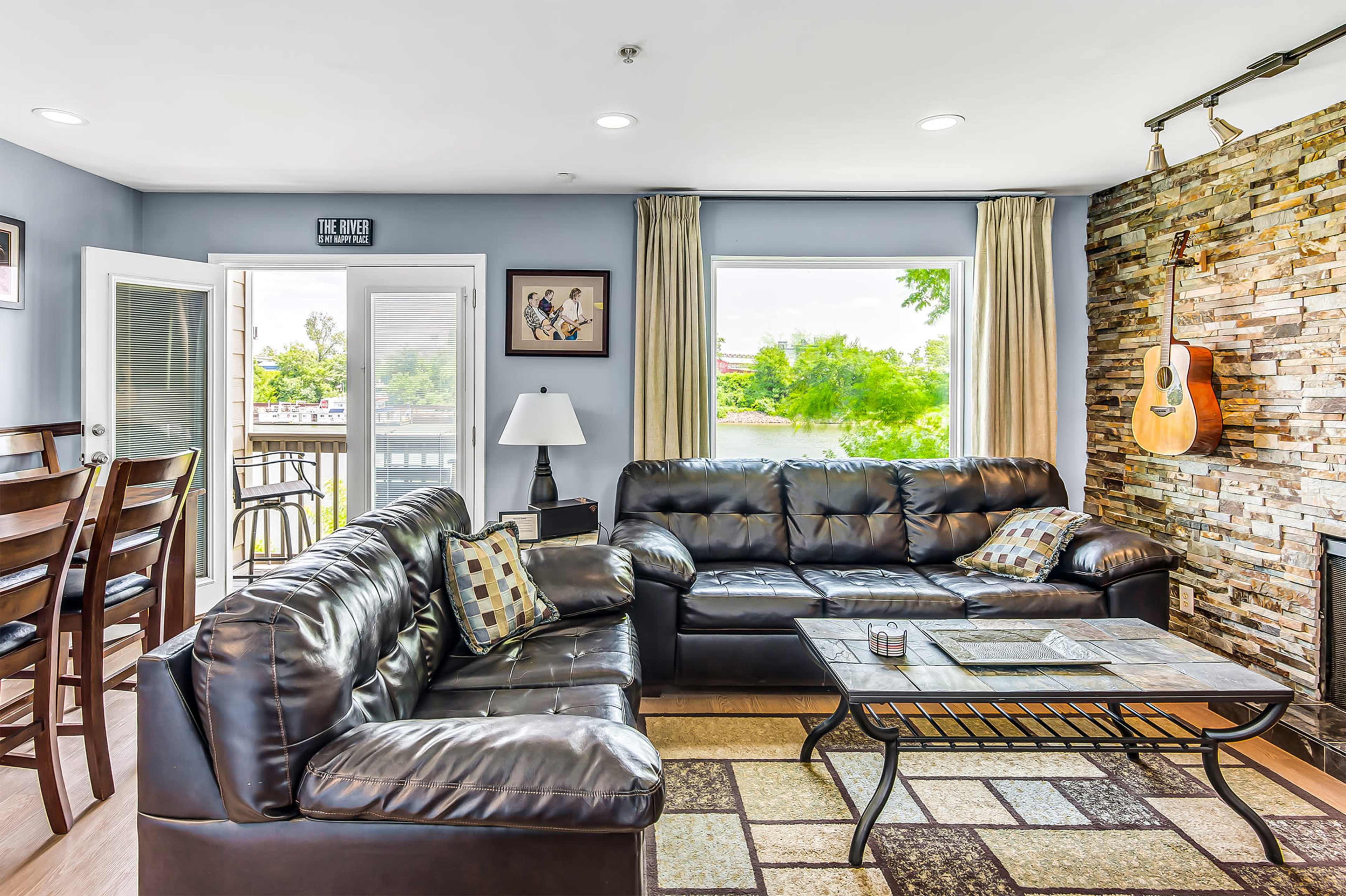 The living room features black leather sofas, a coffee table, and a stone accent wall, with a large window overlooking a scenic view.