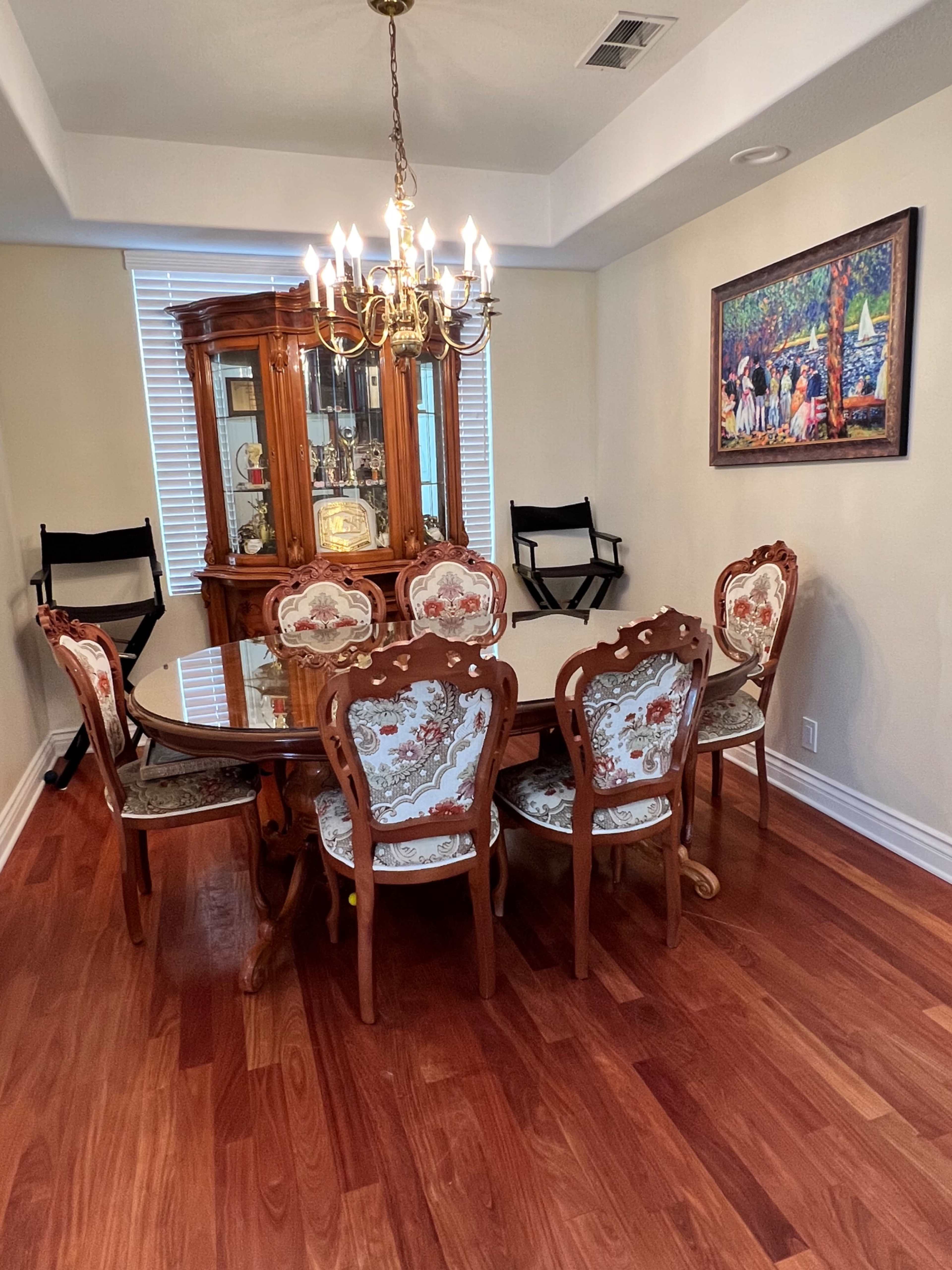 A wooden dining table with ornate chairs is set in a well-lit dining room that features a chandelier and a china cabinet.