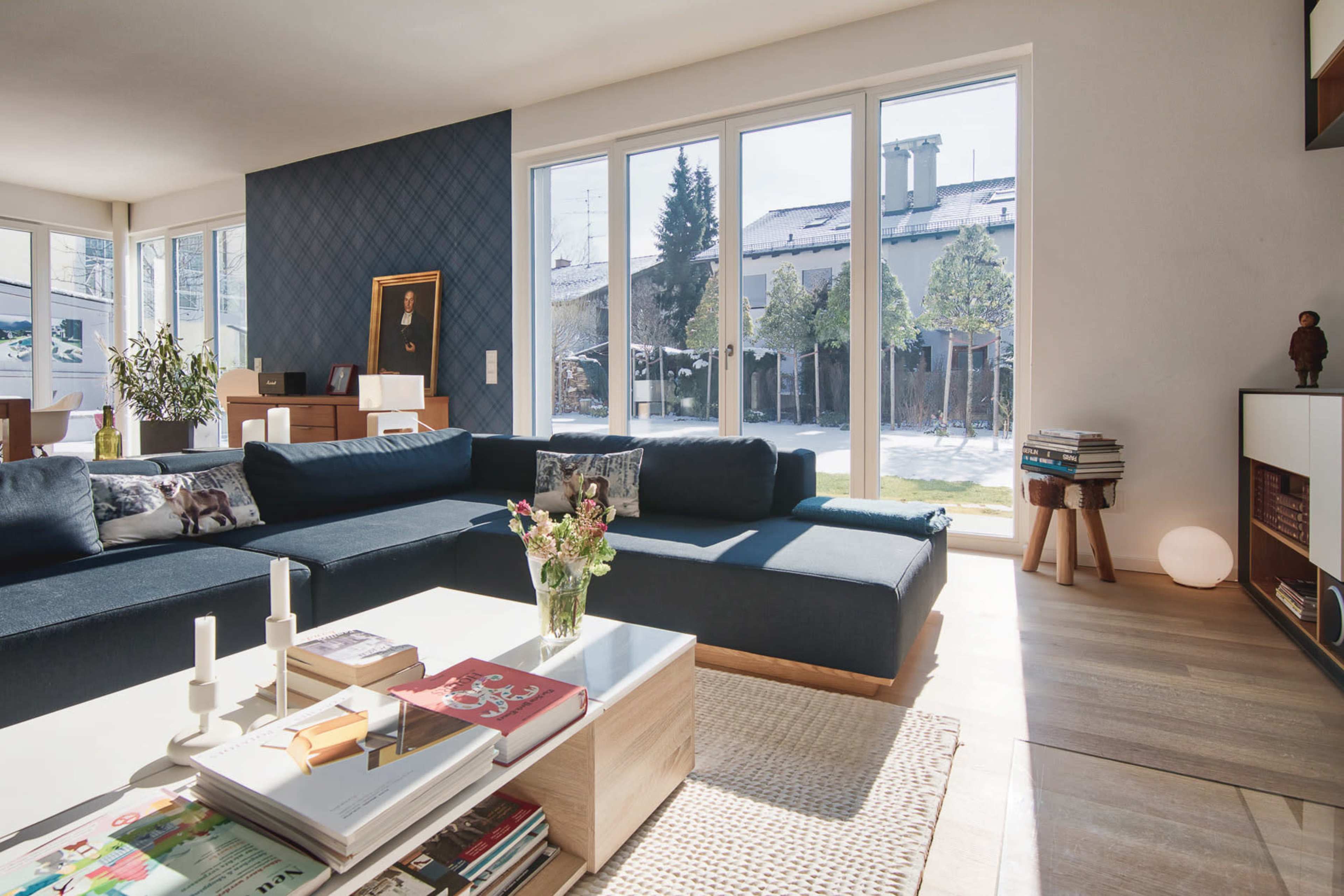 A spacious living room features a dark blue sectional sofa, a glass coffee table with various magazines, and large windows overlooking a snowy outdoor area.