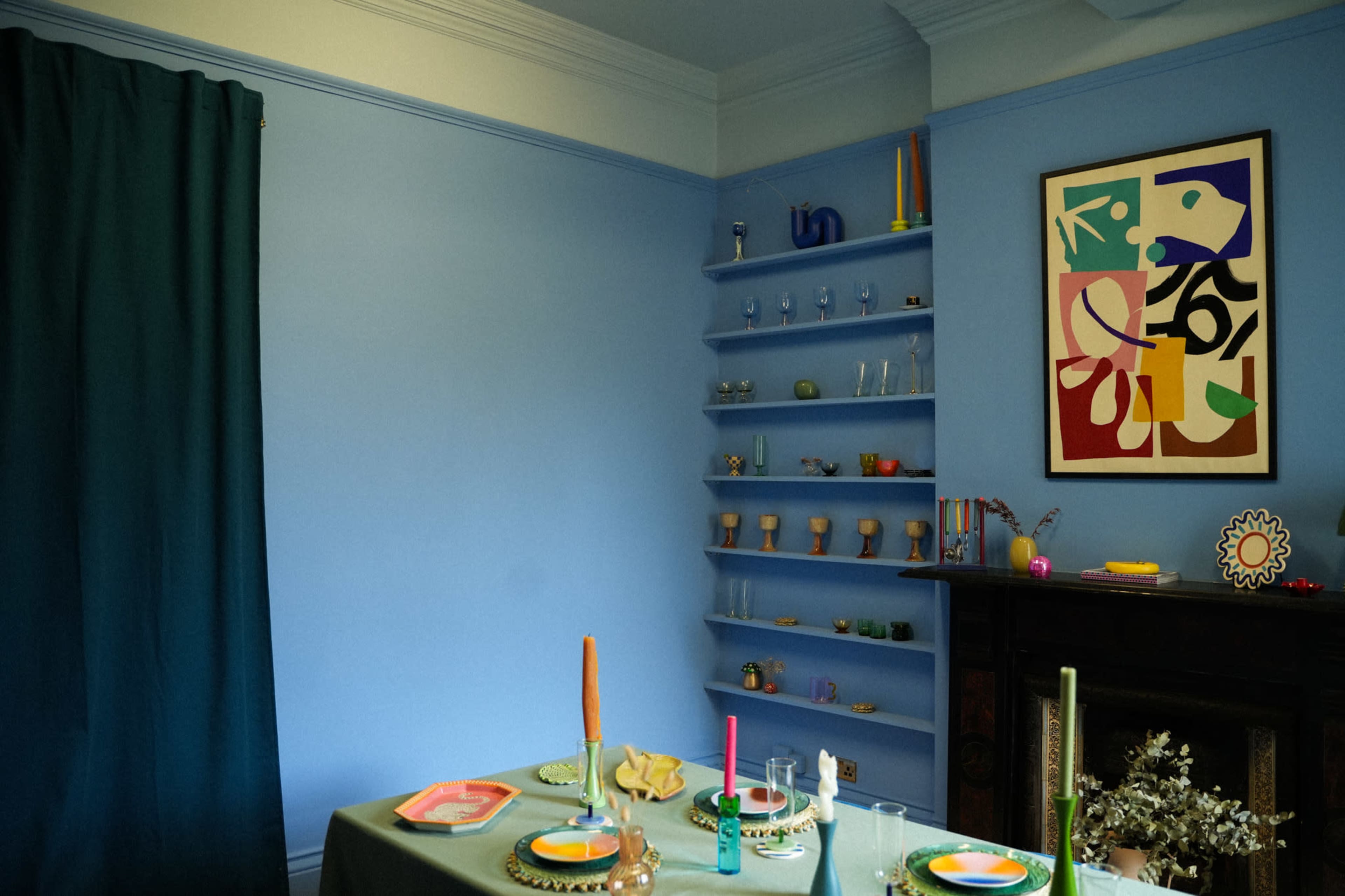 A dining table is set with colorful plates and candles in a room featuring blue walls, a shelf with assorted glassware, and a framed abstract artwork.