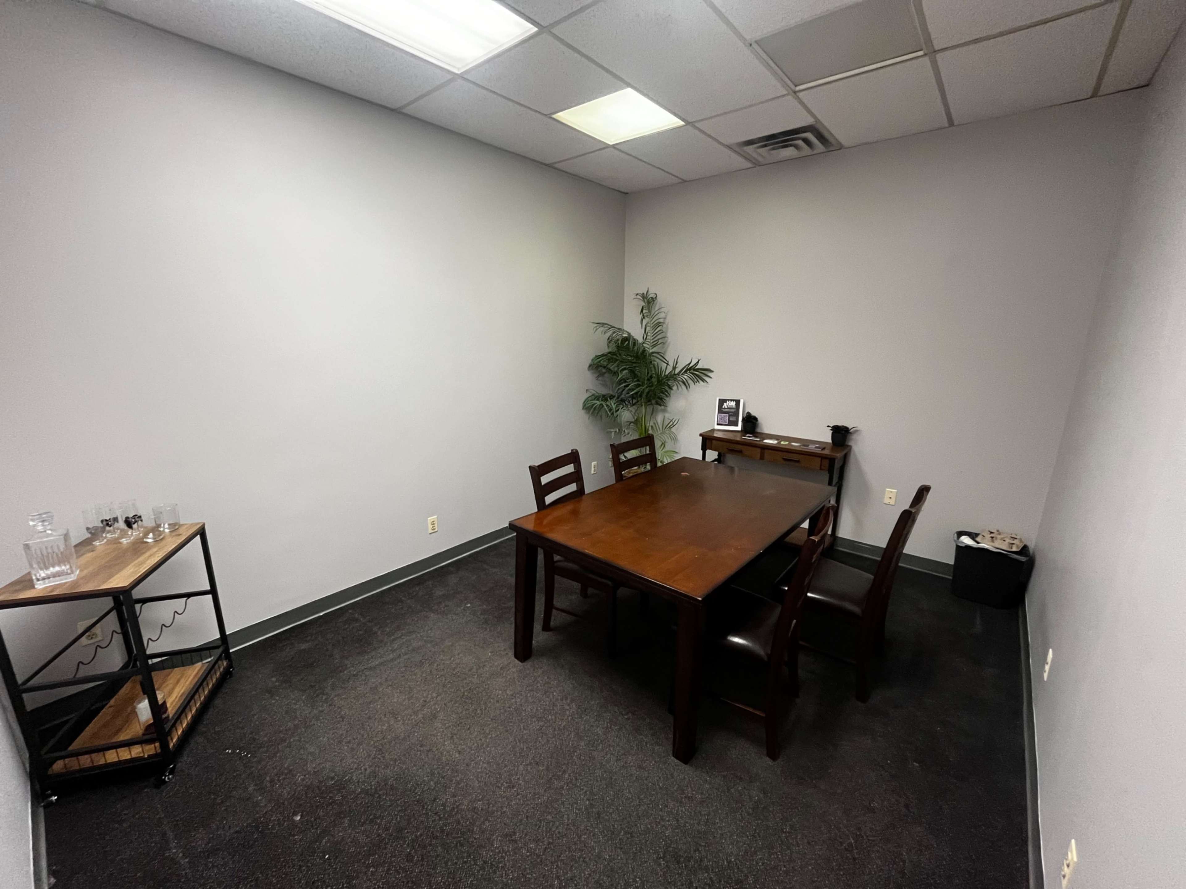 A sparsely furnished meeting room with a wooden table, four chairs, a small shelving unit, and a potted plant in the corner.