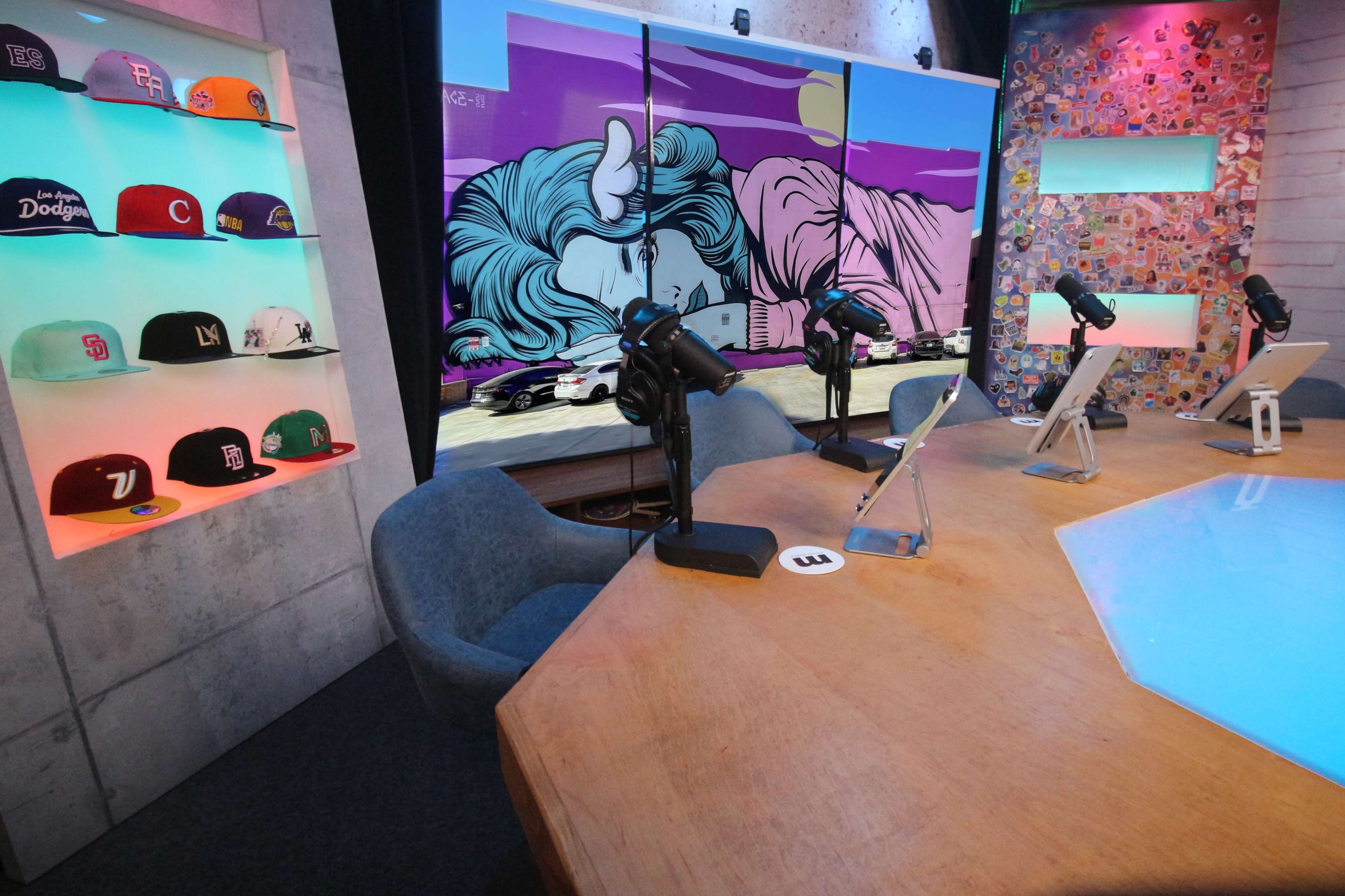 The image shows a modern podcast studio featuring microphones, colorful hats displayed on shelves, a large graphic backdrop, and a wooden table.