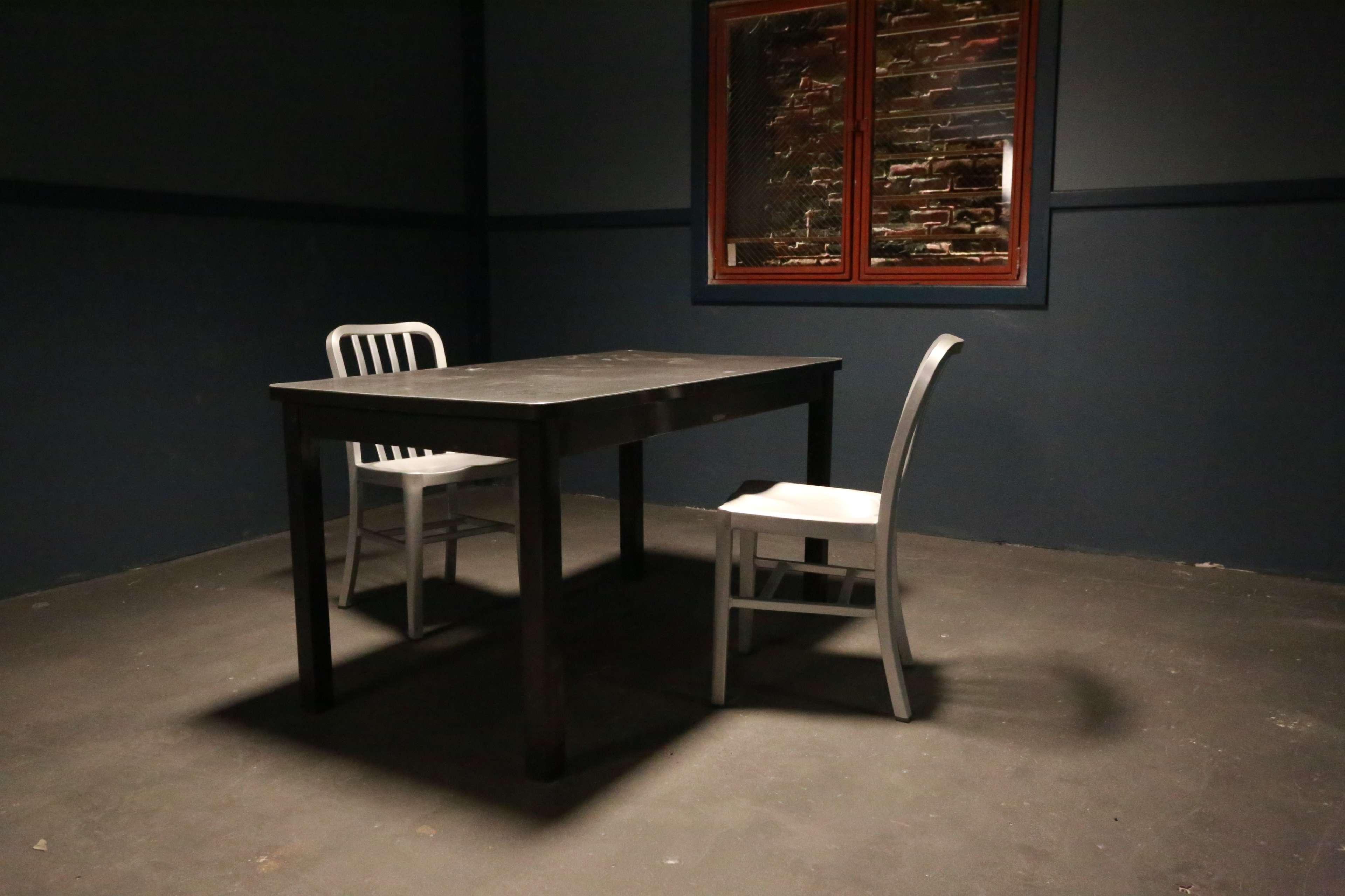 A dark room contains a simple wooden table surrounded by two chairs, with a window showing a brick wall.