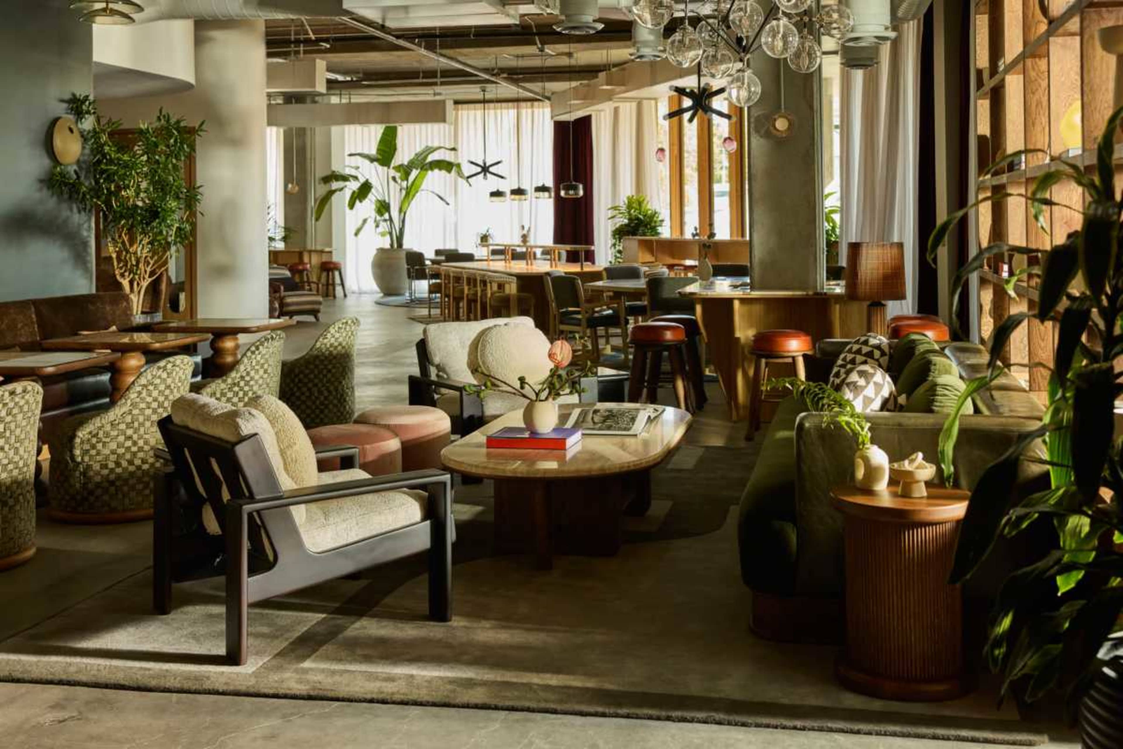 The image shows a modern lounge area with a combination of upholstered seating, wooden tables, plants, and large windows allowing natural light to fill the space.