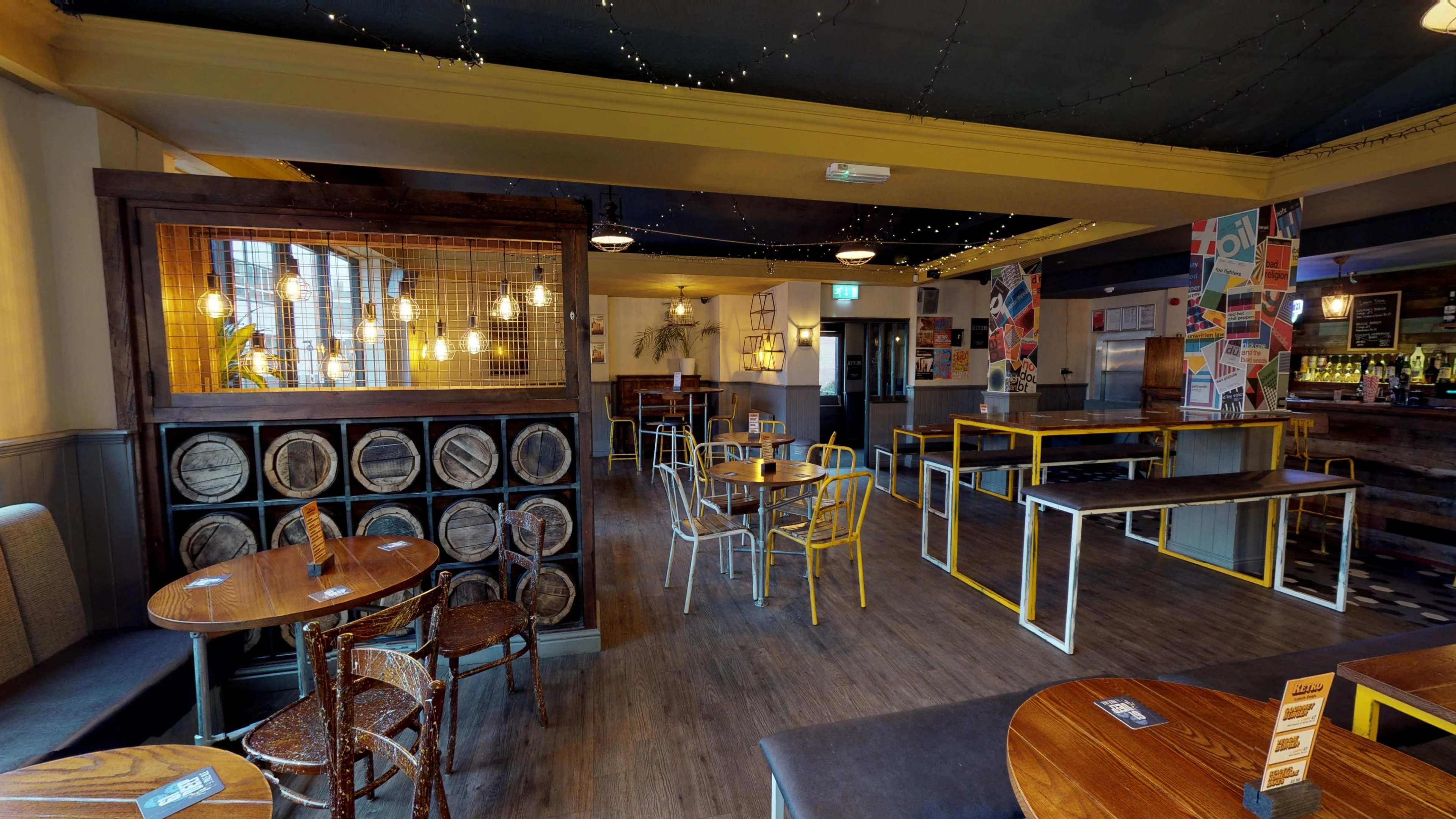 The image shows a spacious bar interior with wooden and metal furniture, featuring a mix of tables and chairs, illuminated by warm pendant lights.