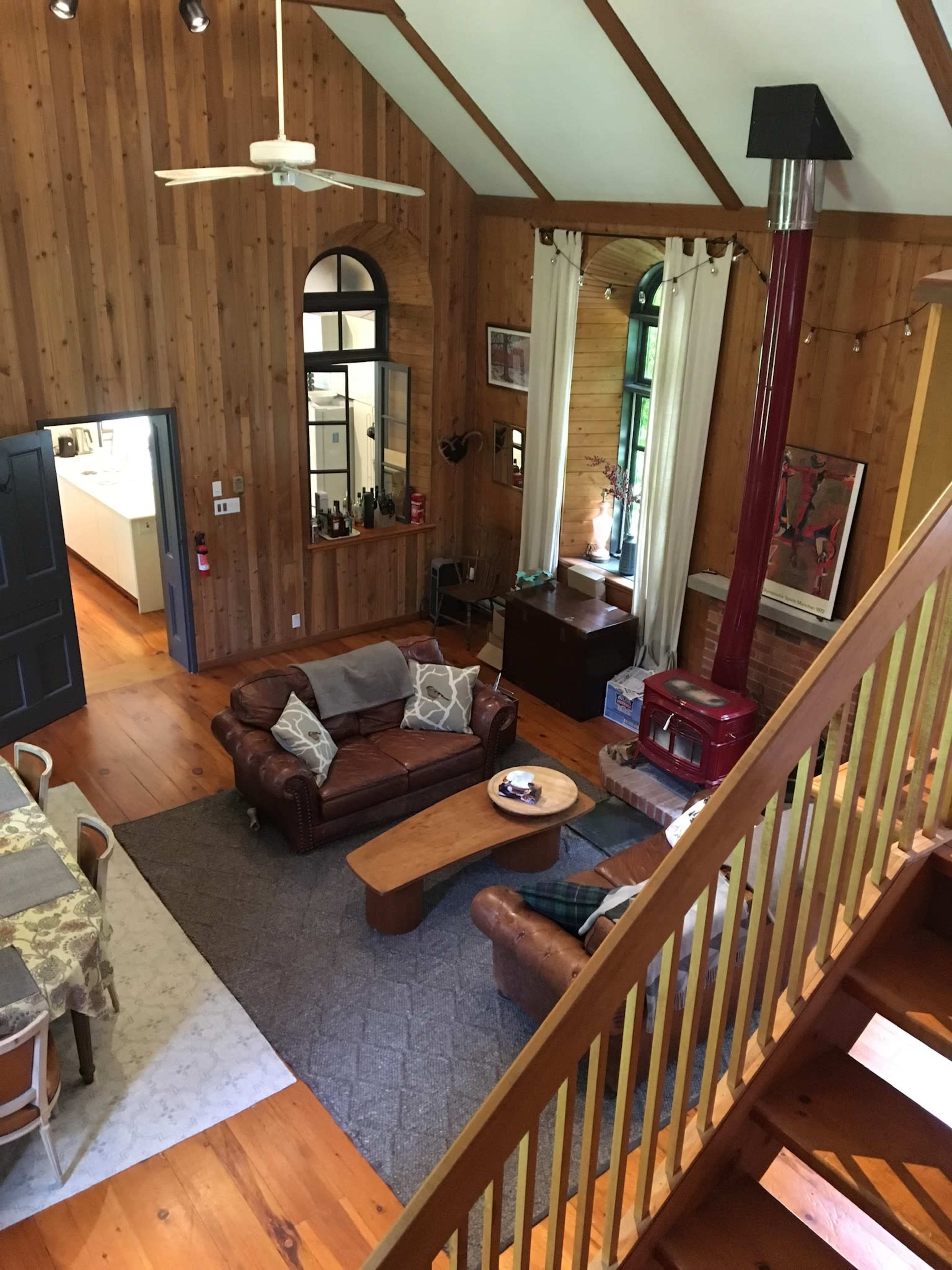 A spacious living room with wooden walls, a large leather sofa, a coffee table, and a red stove, viewed from an upper level.