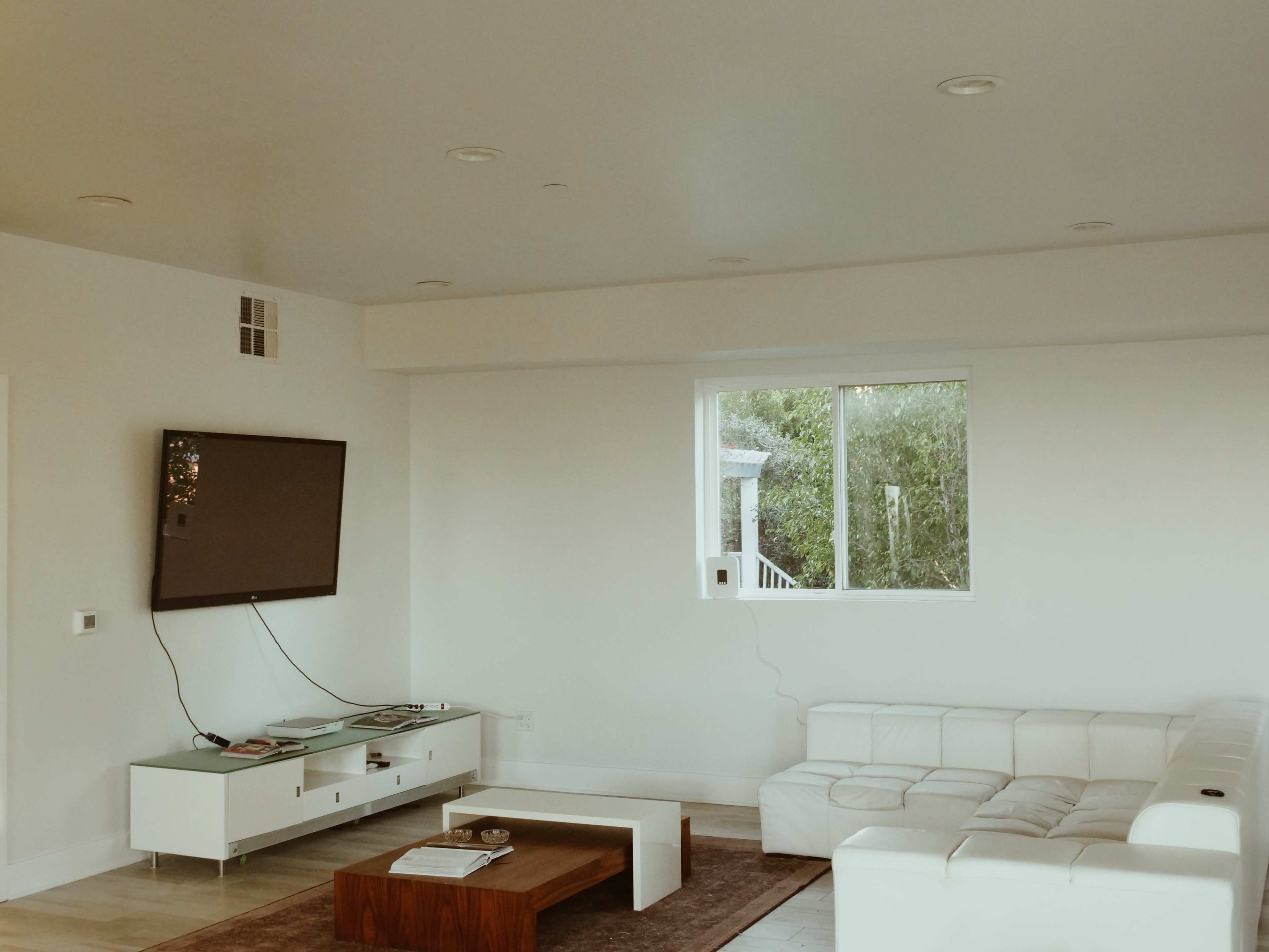 The image shows a modern living room with a white sofa, a wall-mounted television, and a large window overlooking greenery.