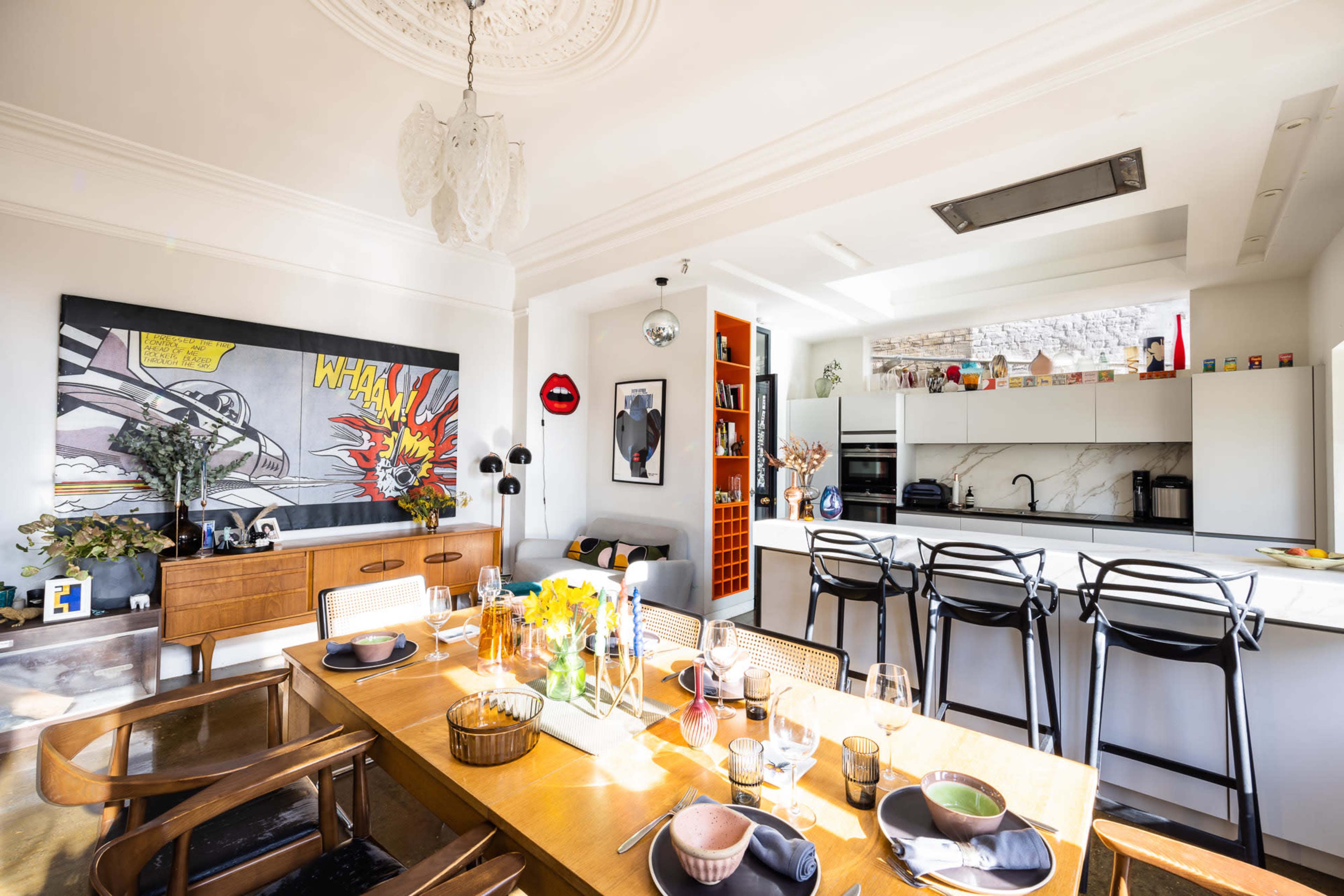 The image shows a modern kitchen and dining area with a large table set for a meal, featuring a vibrant pop art piece on the wall.