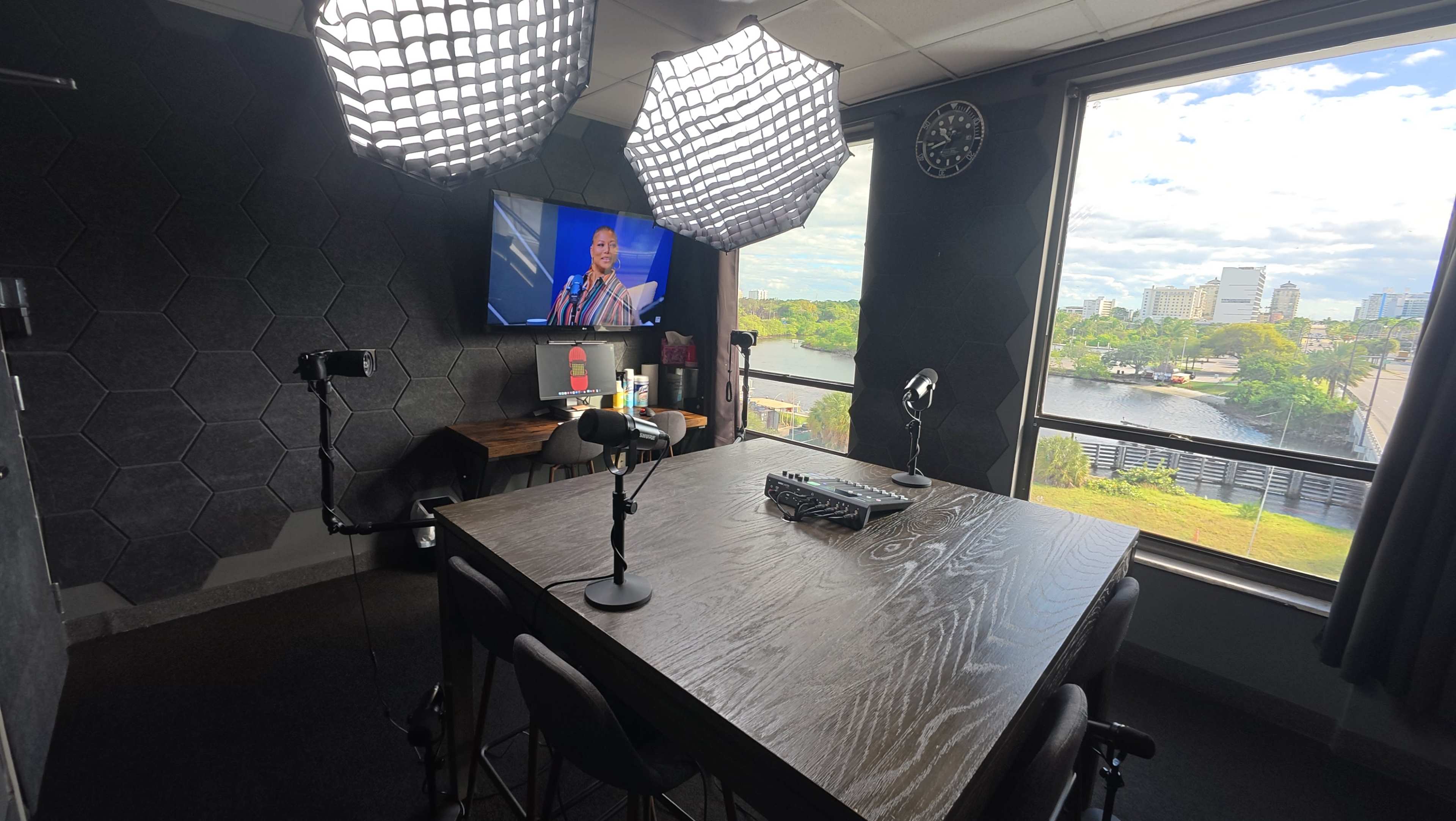 The image shows a modern workspace with a large wooden table, two light setups, a television displaying a person, and a view of a cityscape through a window.