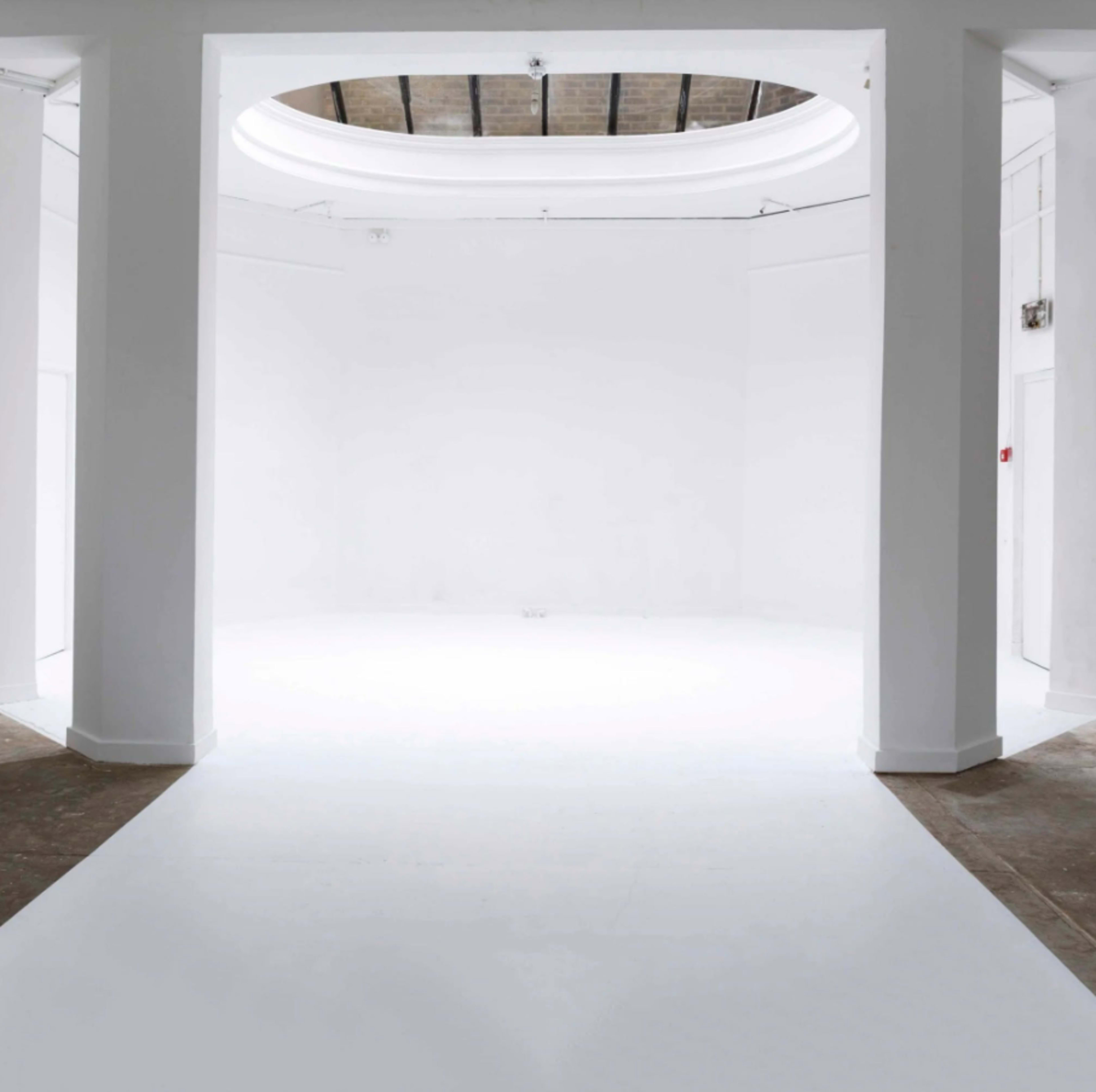 The image shows a spacious, empty room with white walls and a white floor, featuring a circular ceiling opening and pillars.