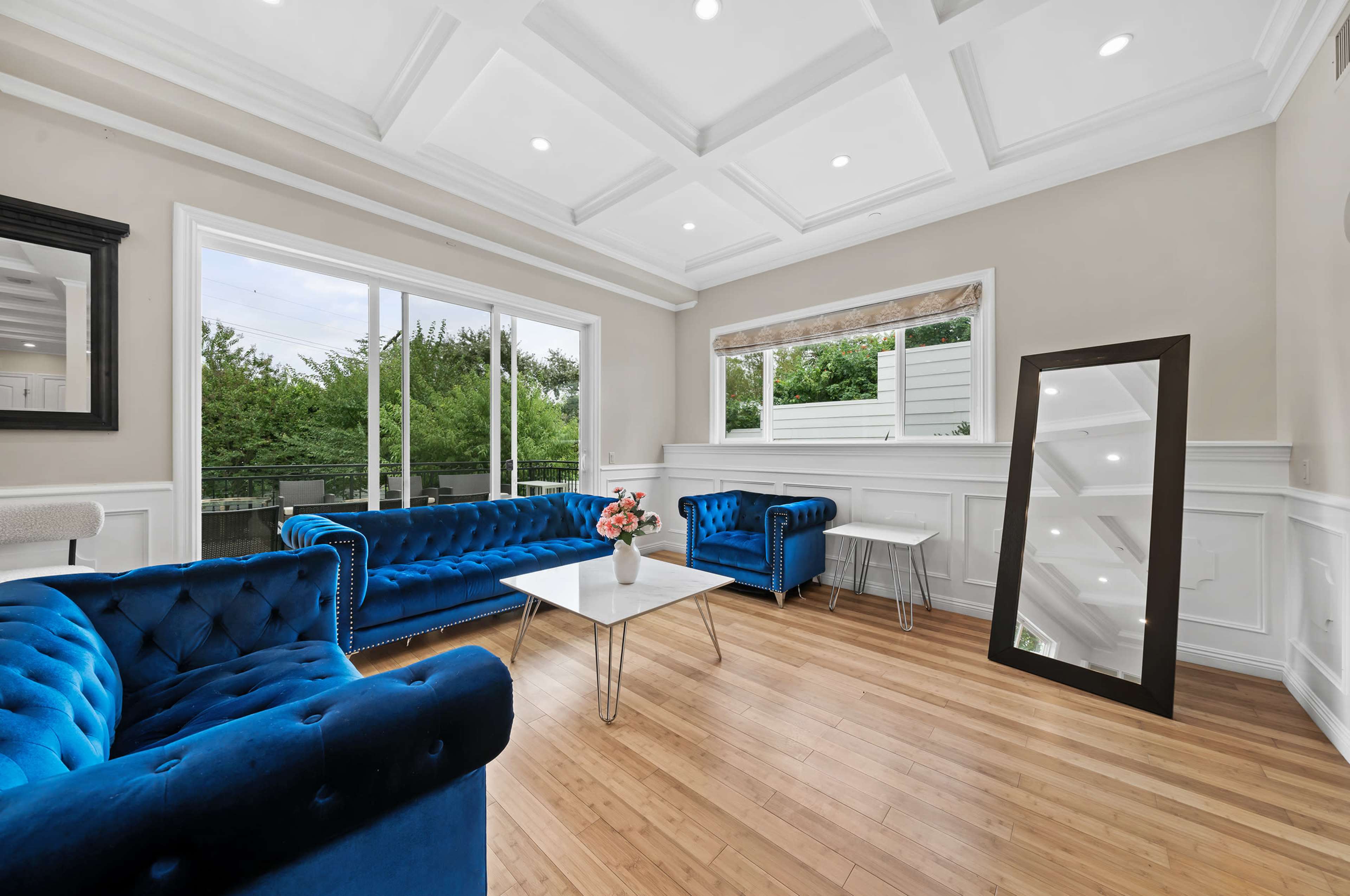 The image shows a bright living room with blue tufted sofas, a glass coffee table, and a large mirror against a wall with white paneling.