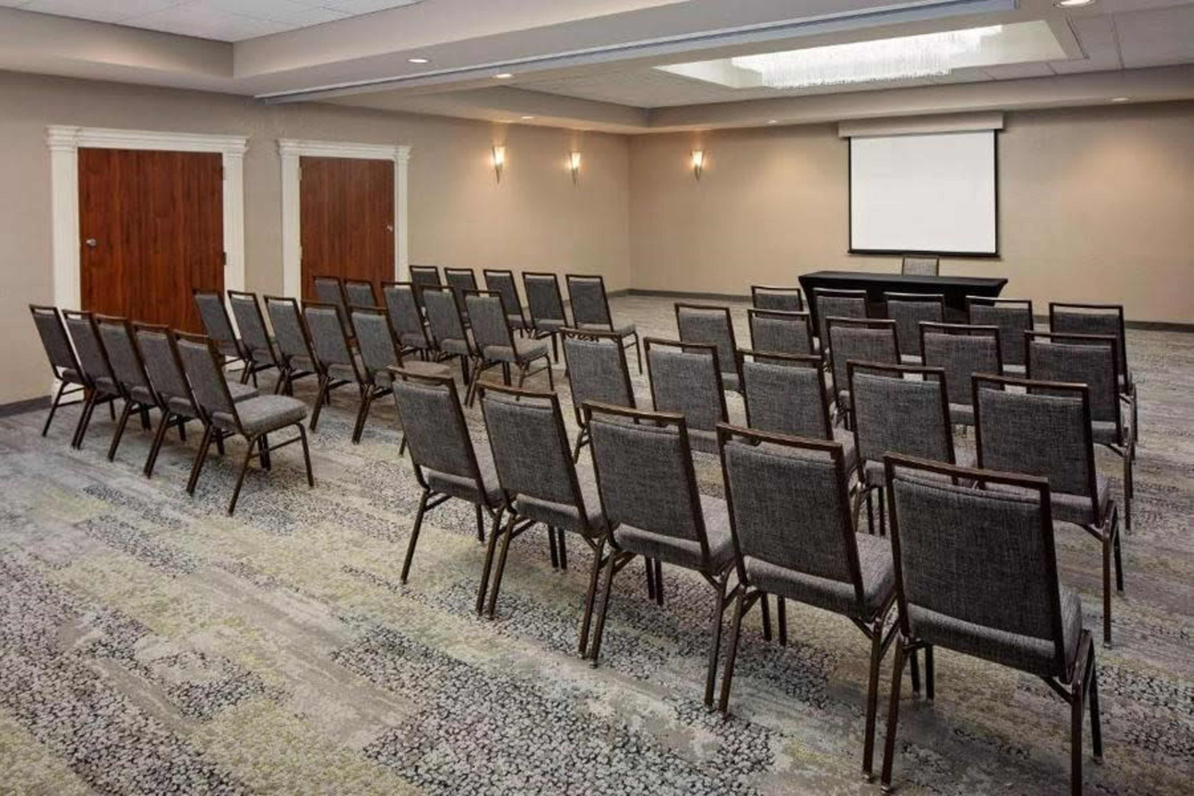 The image shows a conference room arranged with rows of gray chairs facing a screen at the front.
