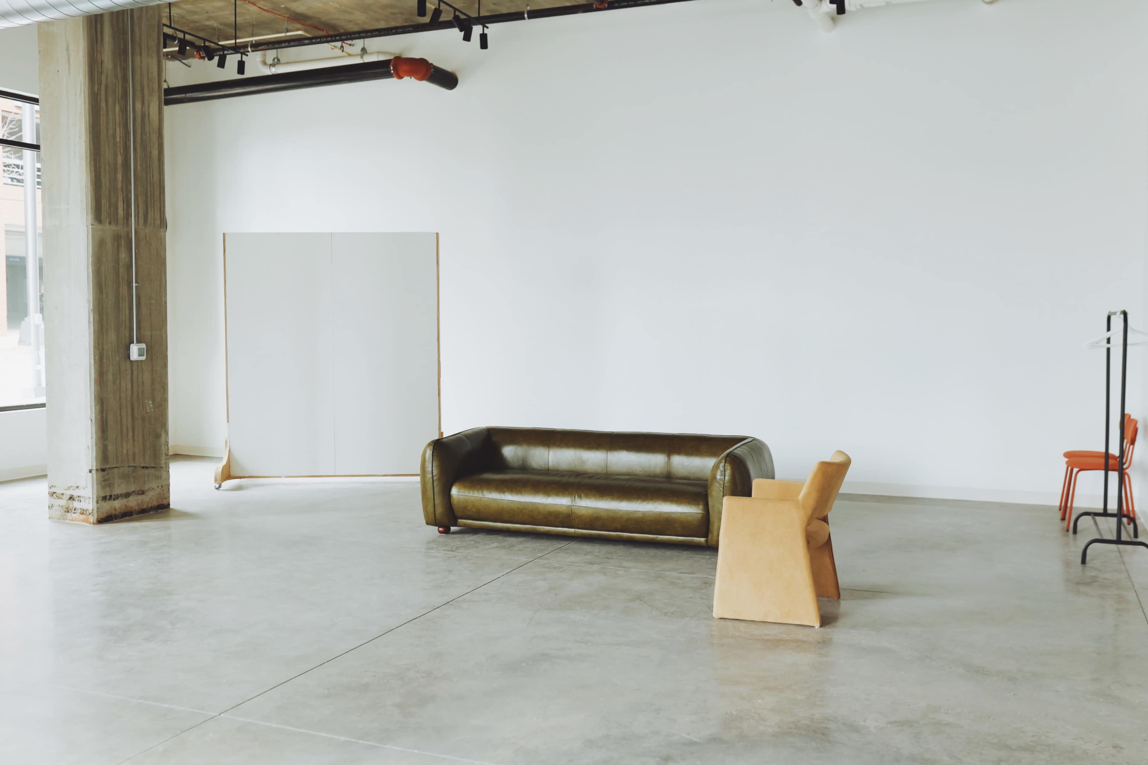 A spacious, minimalist room with a green sofa, a wooden chair, and a white wall with a large canvas propped against it.