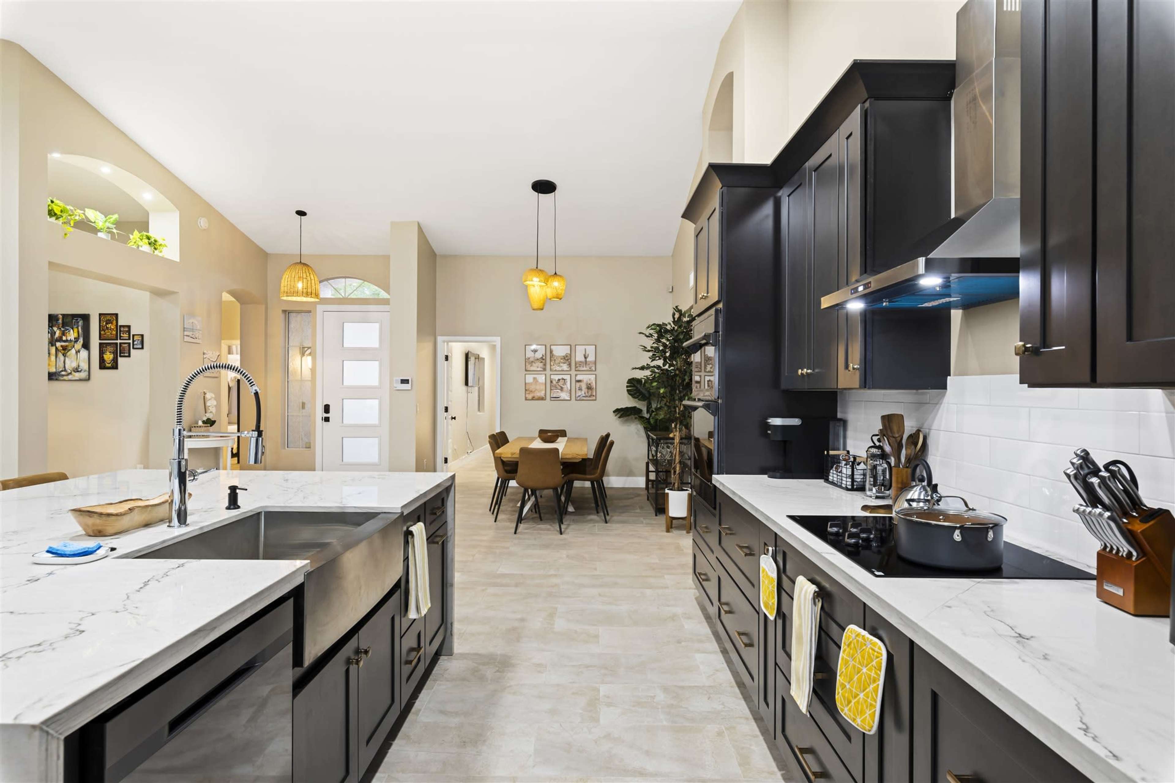 A modern kitchen with dark cabinetry, a large island with a stainless steel sink, and a bright dining area in the background.