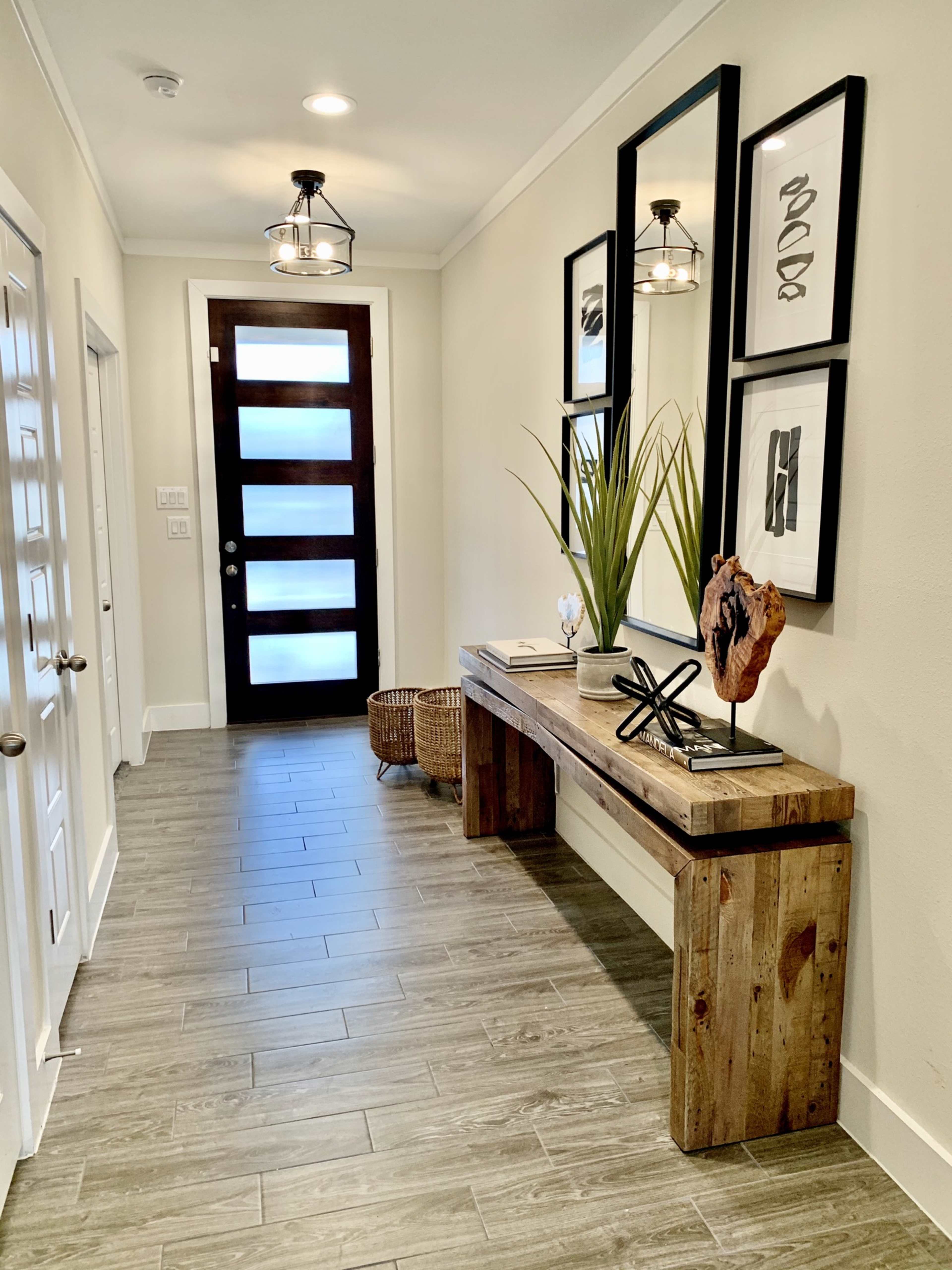 A modern hallway featuring a wooden console table, a decorative plant, framed artwork on the walls, and a large front door with multiple glass panels.