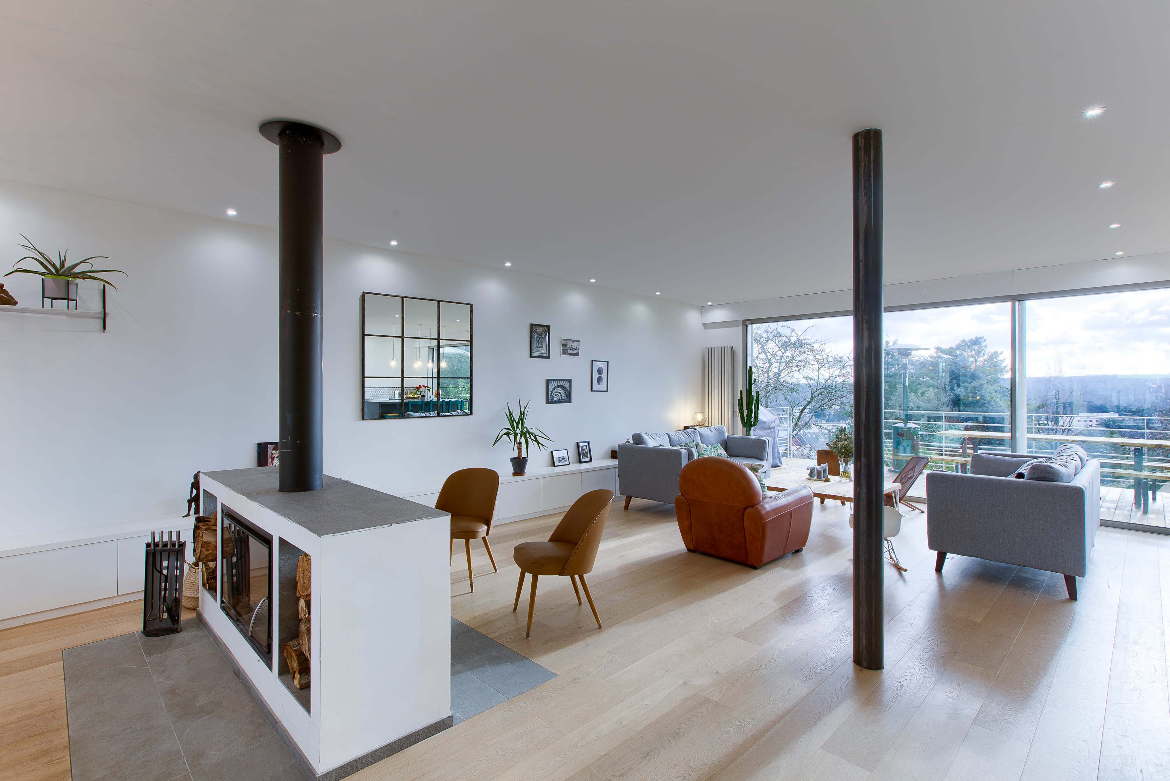 A modern living room features a central fireplace, large windows offering a view, and a mix of comfortable seating arrangements.
