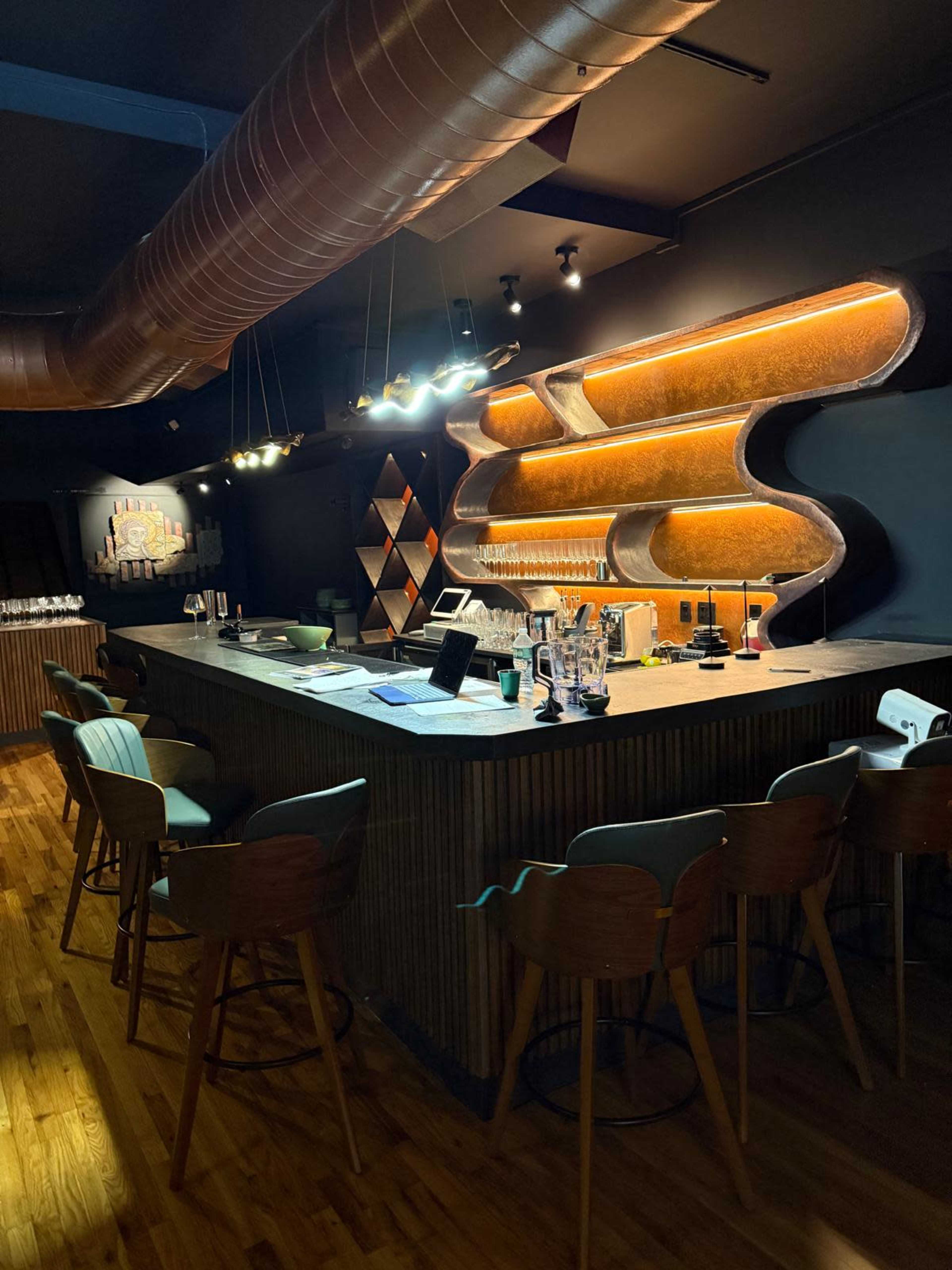 The image shows a modern bar area with a sleek counter, illuminated shelves, and a mix of wooden and upholstered seating.
