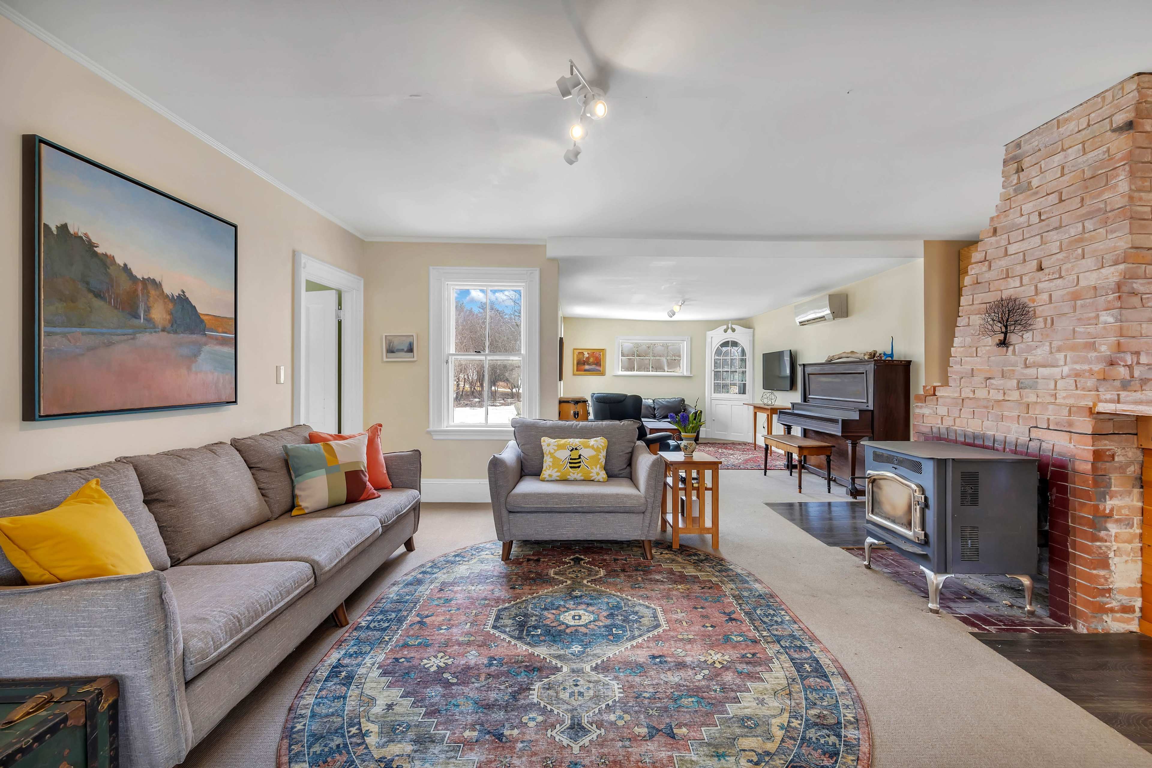 A spacious living room features a cozy sofa, a patterned rug, a wood-burning stove, and a piano, with large windows allowing natural light to enter.