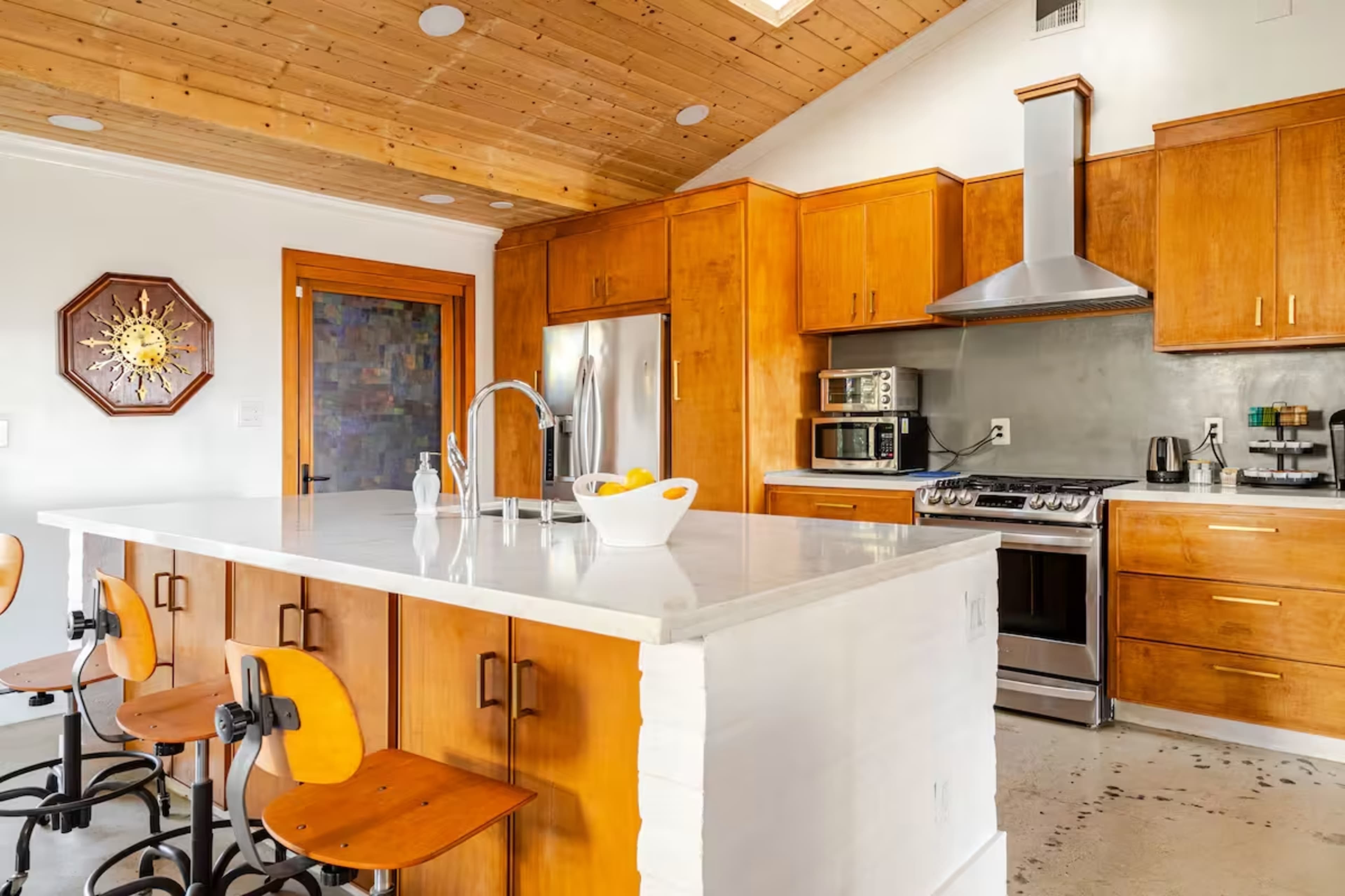 The image shows a modern kitchen with wooden cabinets, a large island with seating, stainless steel appliances, and a vaulted ceiling with wooden beams.
