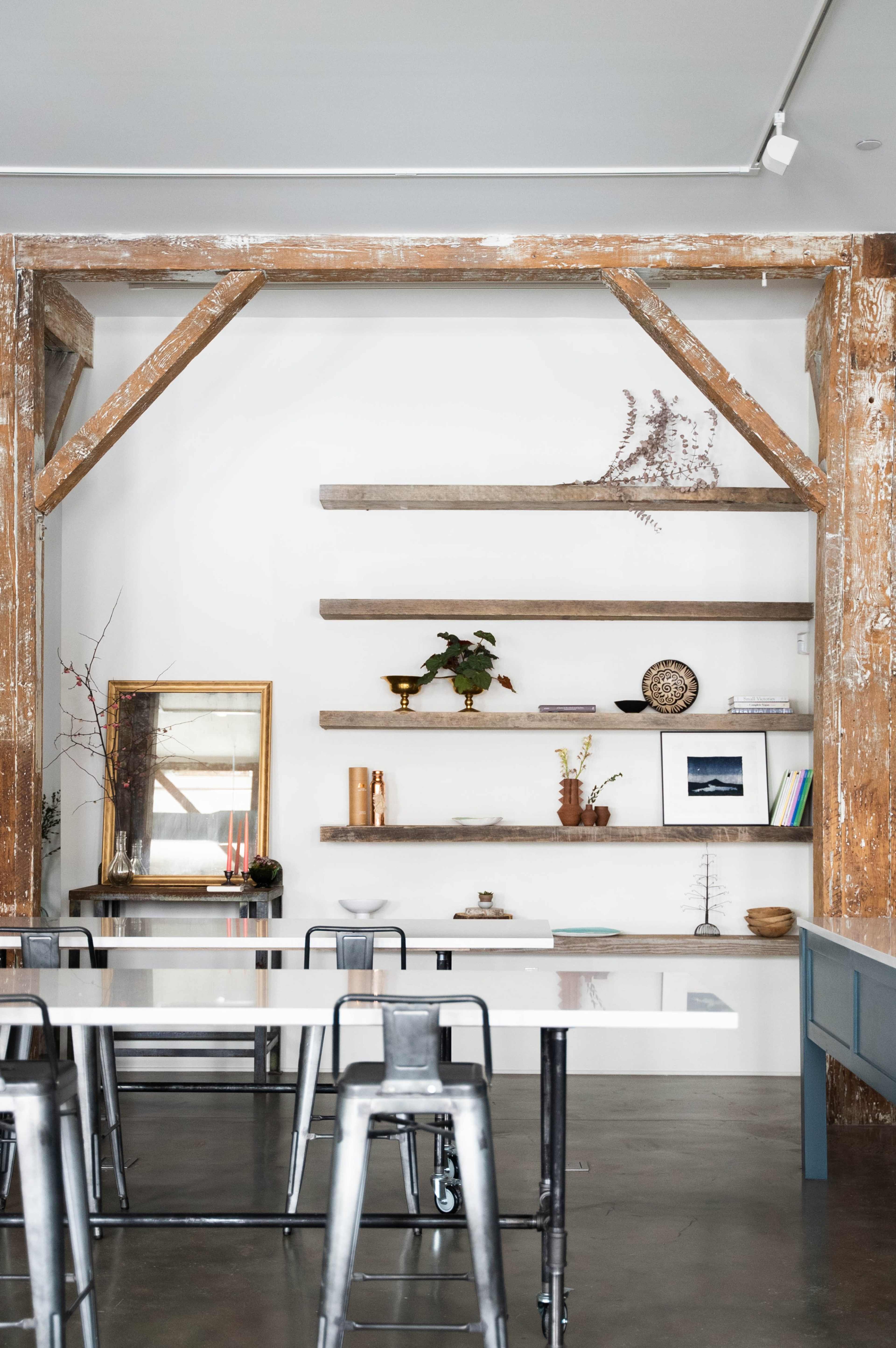 A modern interior of a café featuring wooden shelves mounted on a white wall, with a mix of decorative items and plants, along with metal tables and chairs arranged beneath.