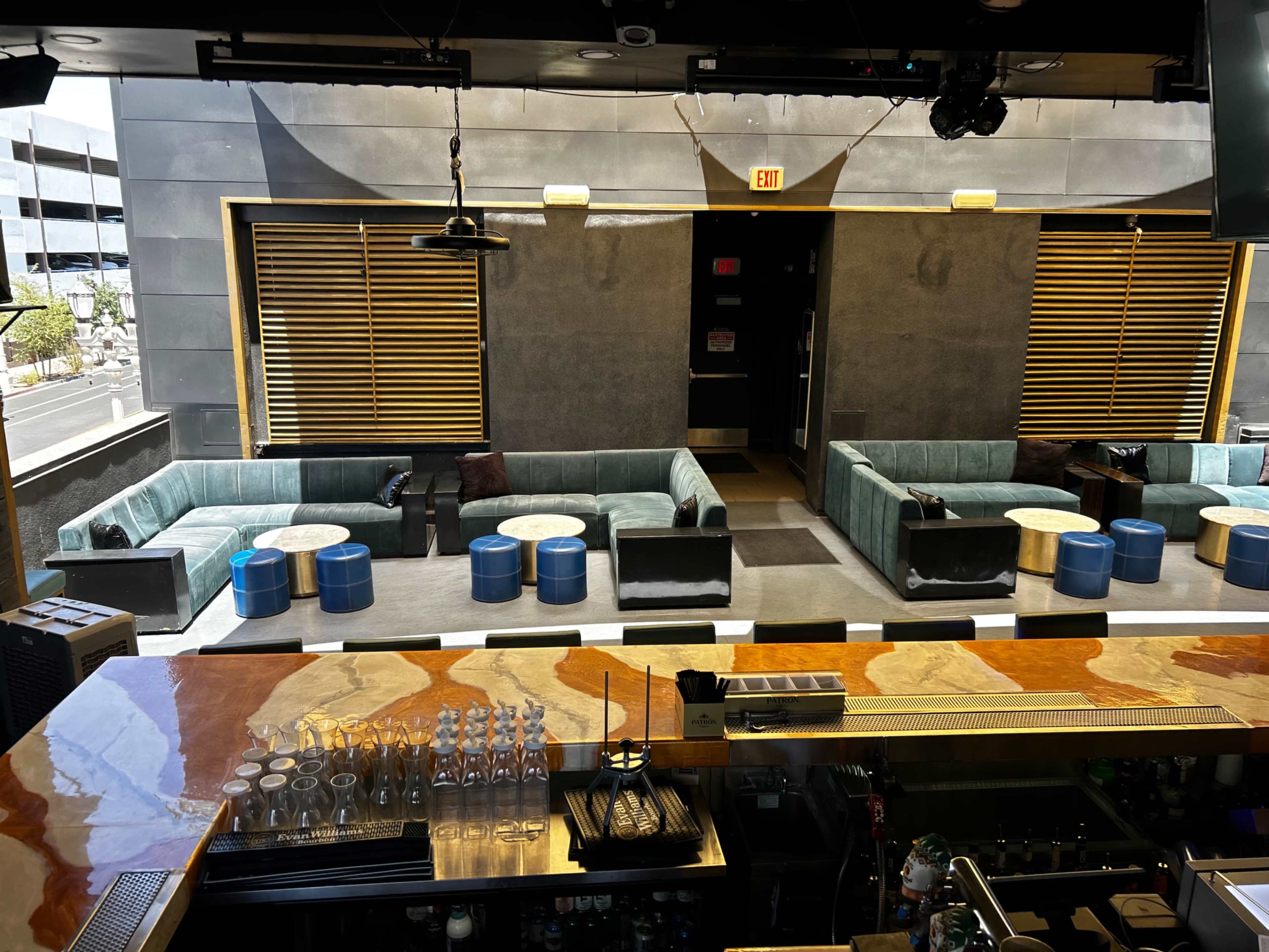 The image shows a bar interior with a wooden countertop overlooking an outdoor seating area featuring blue stools and small tables, surrounded by large, modern lounge sofas.