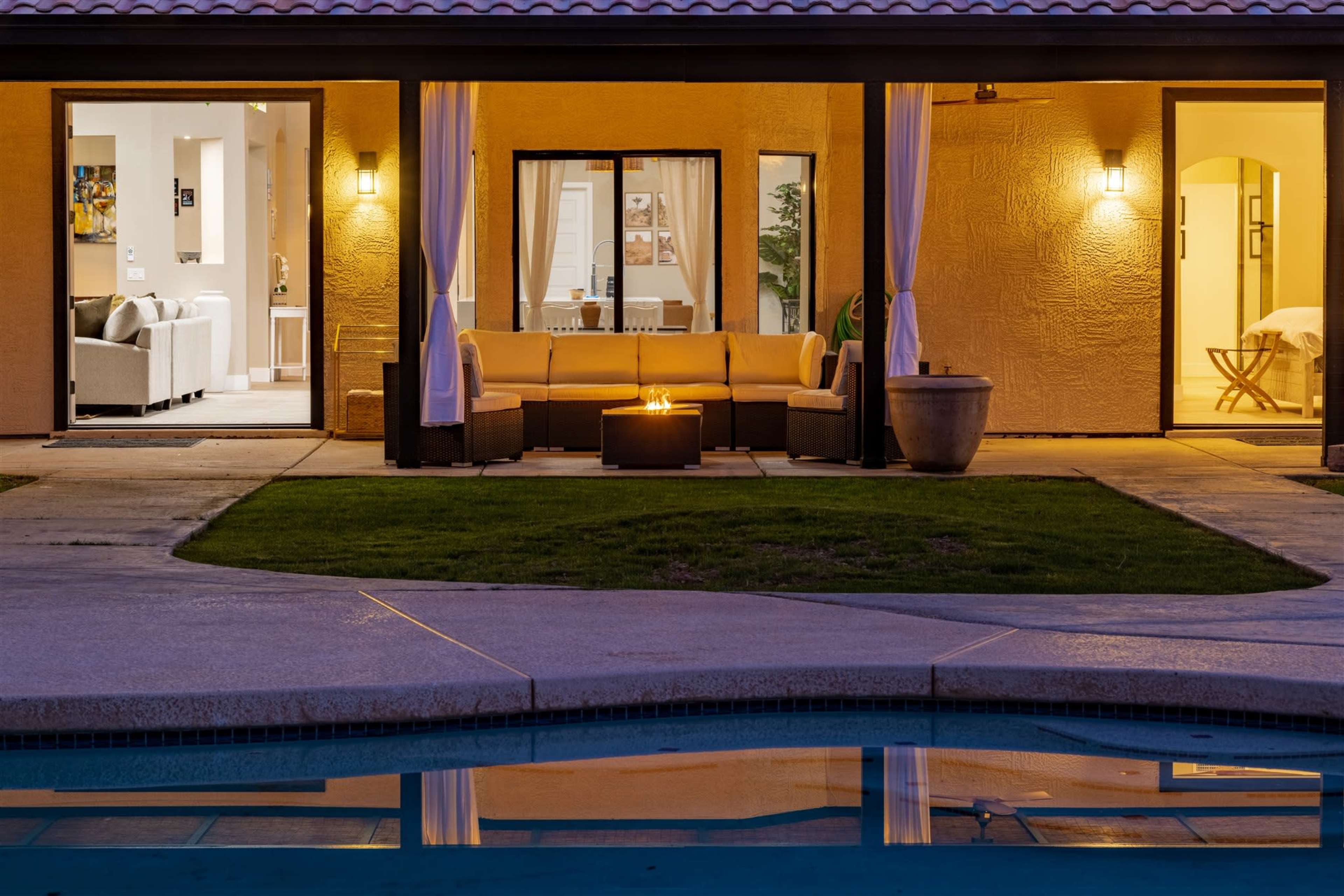 A modern outdoor lounge area with a fire pit is situated beside a swimming pool, reflecting the lights of the adjacent indoor living space.