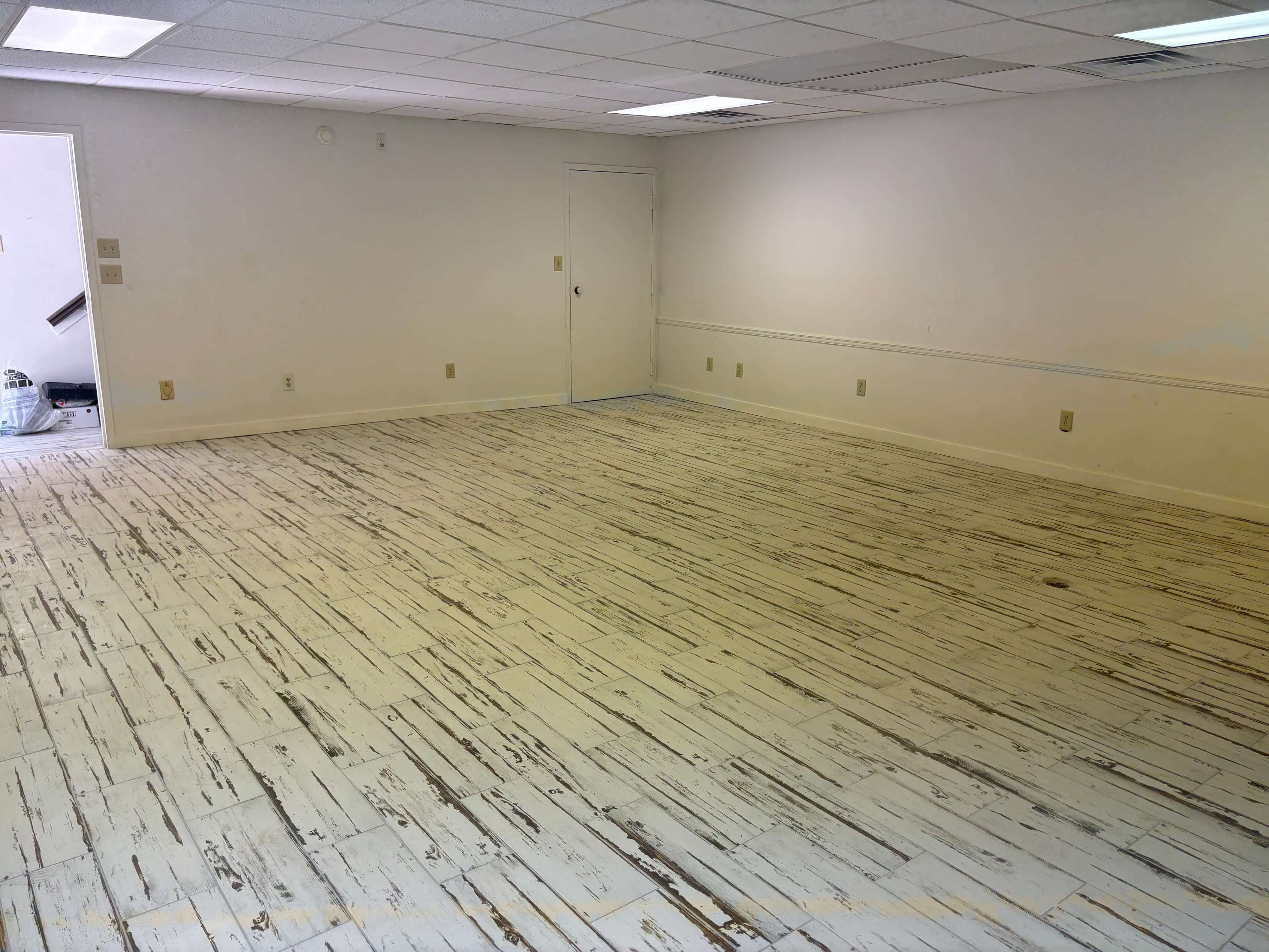 The image shows a spacious, empty room with a weathered wooden floor and white walls, featuring a door on the left side.