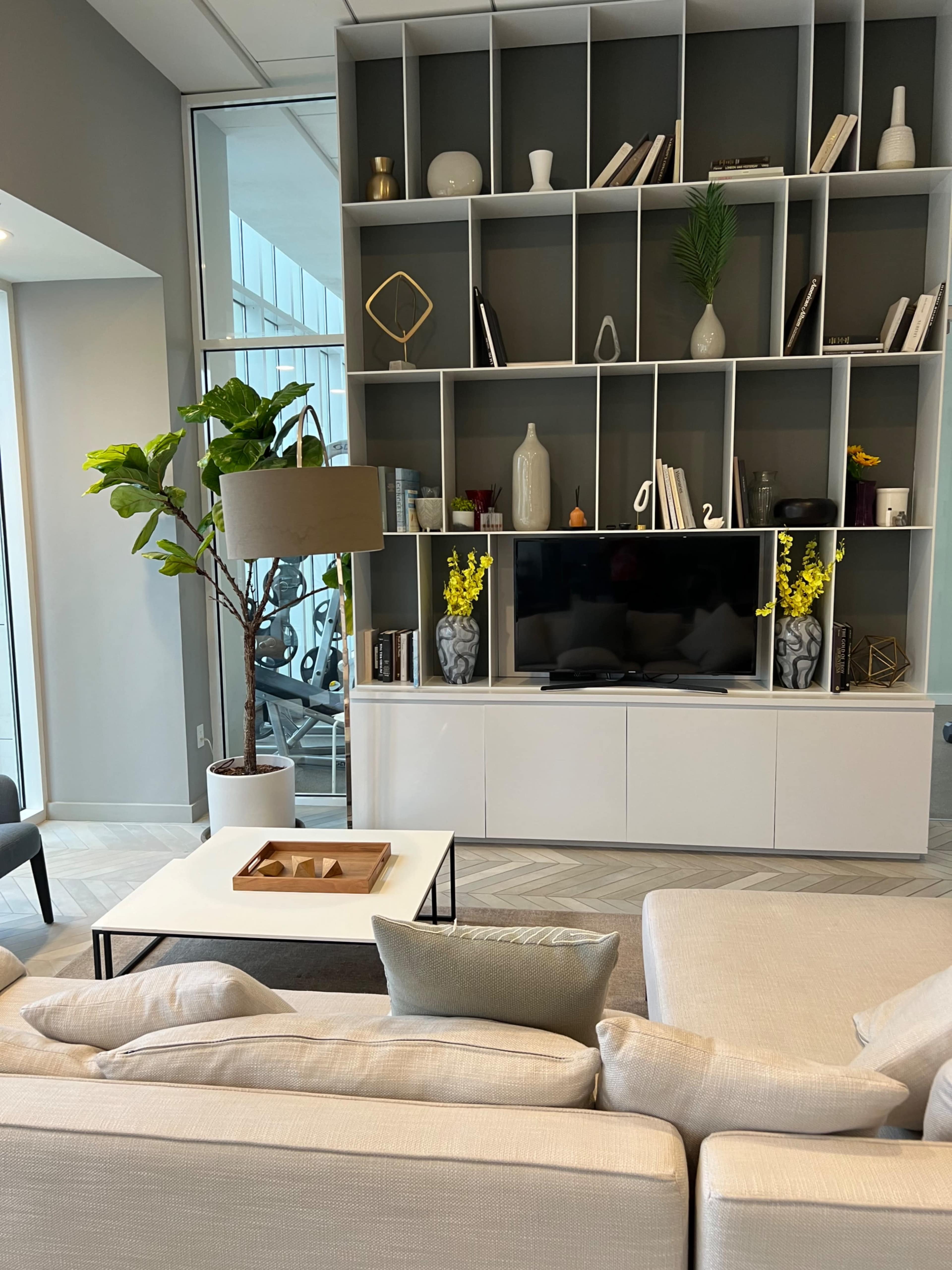 A modern living room features a large bookshelf with decorative items, a television unit, a coffee table, and a plant beside a light-colored sectional sofa.