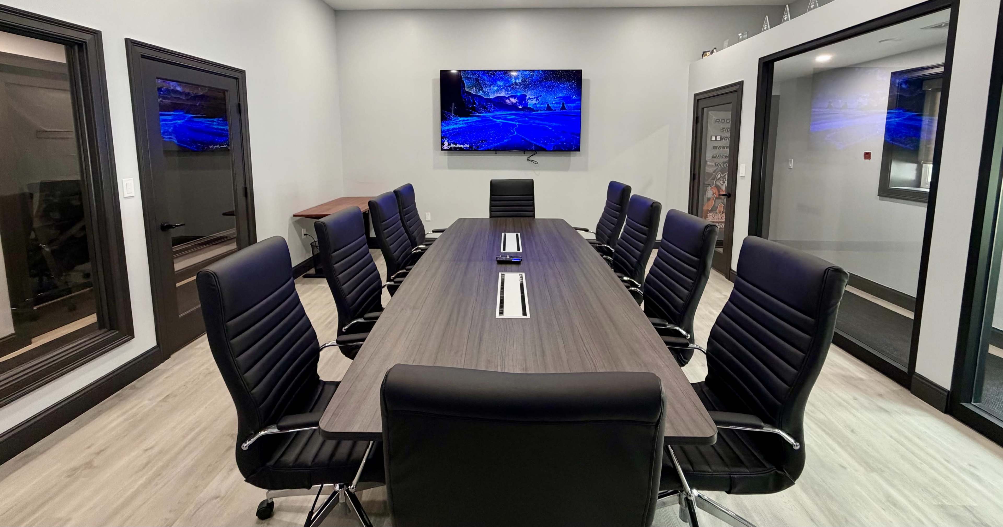 The image shows a modern conference room featuring a long table surrounded by black leather chairs, with a large screen mounted on the wall.