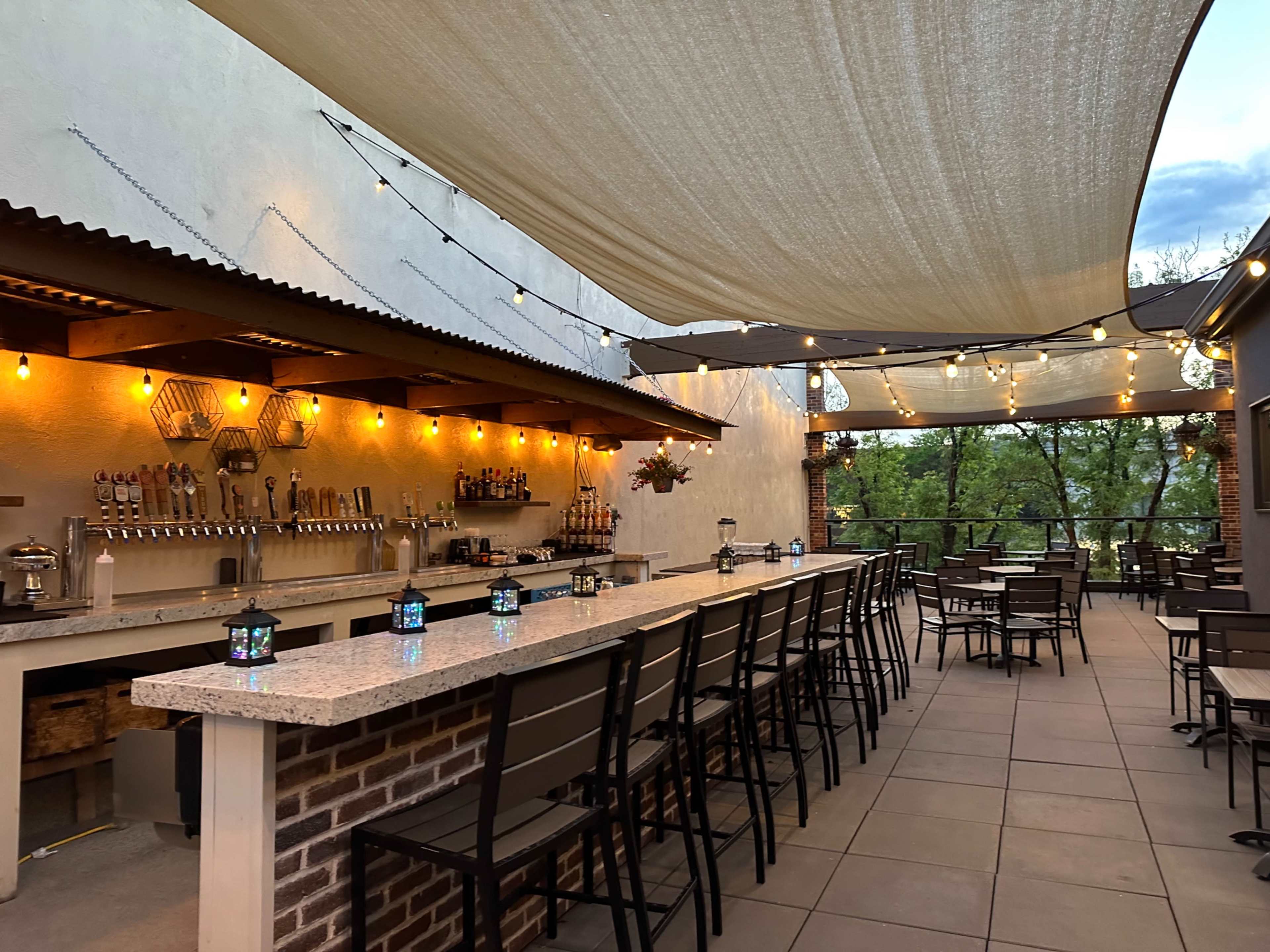 The image shows a modern outdoor bar area with a long counter, rows of chairs, and string lights overhead, set against a backdrop of trees.