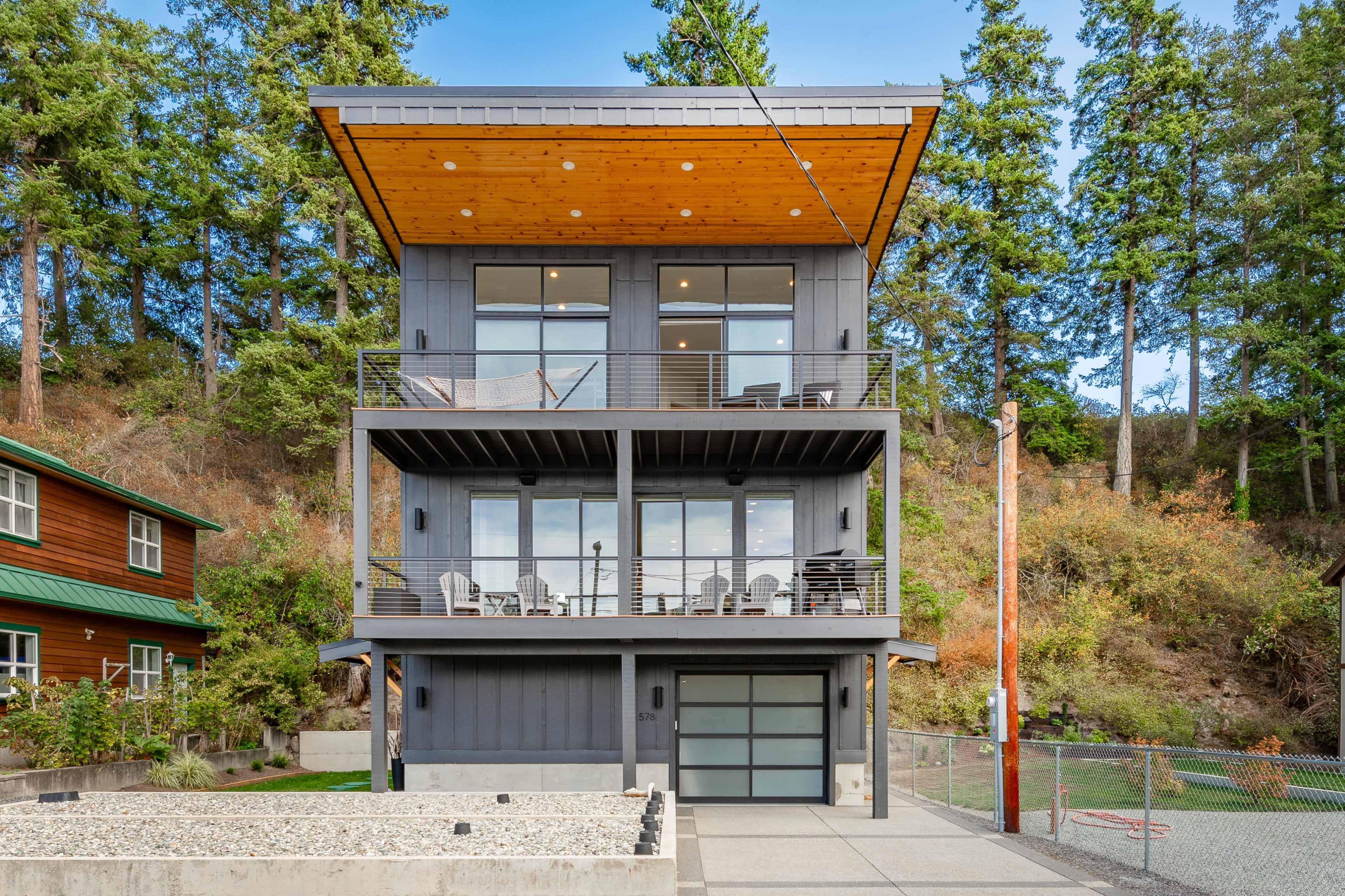 The image shows a modern three-story house with large windows, a wooden overhang, and a concrete driveway, surrounded by trees.