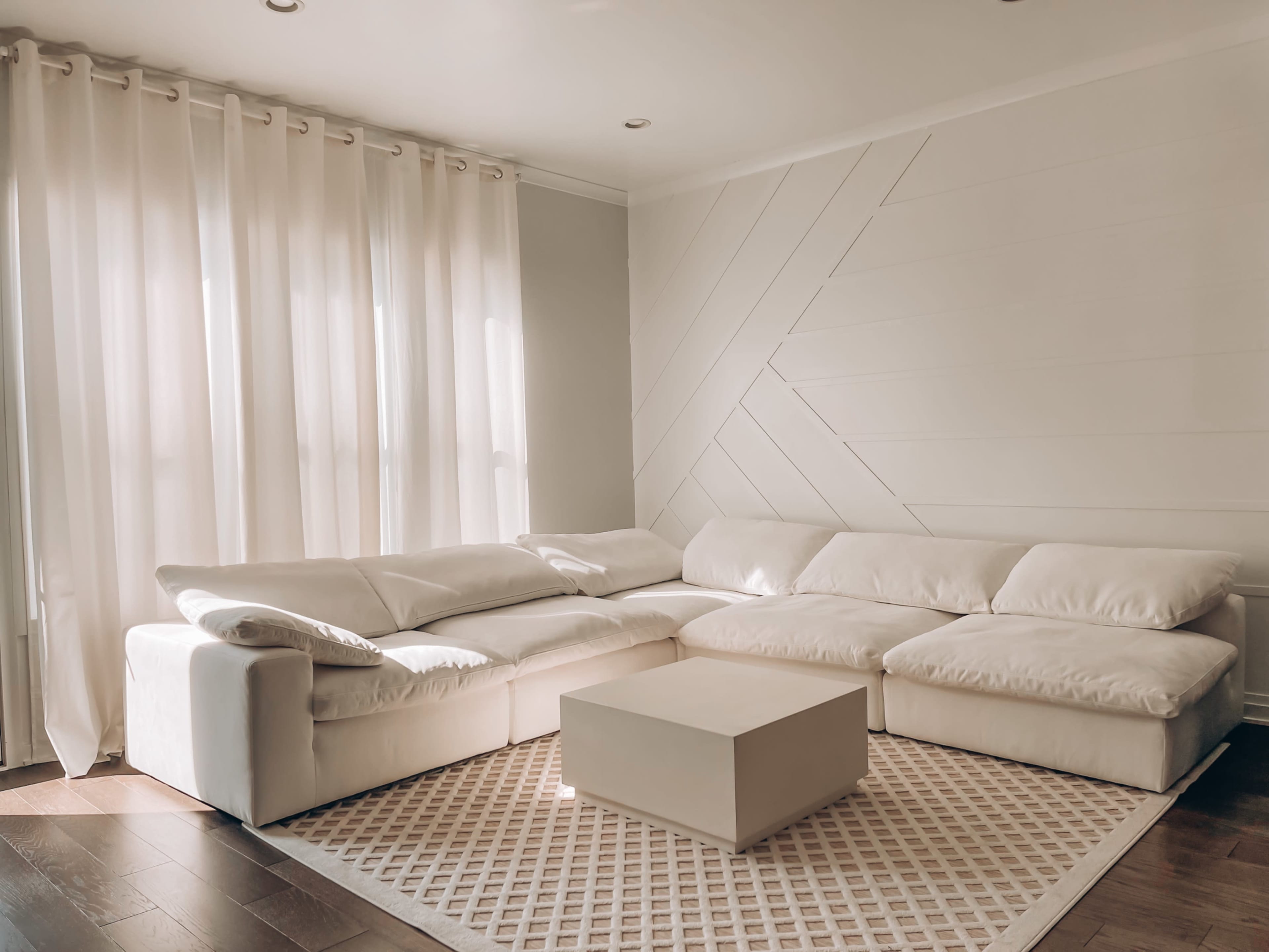 A spacious living room features a white sectional sofa, a light-colored coffee table, and large windows covered with sheer curtains.