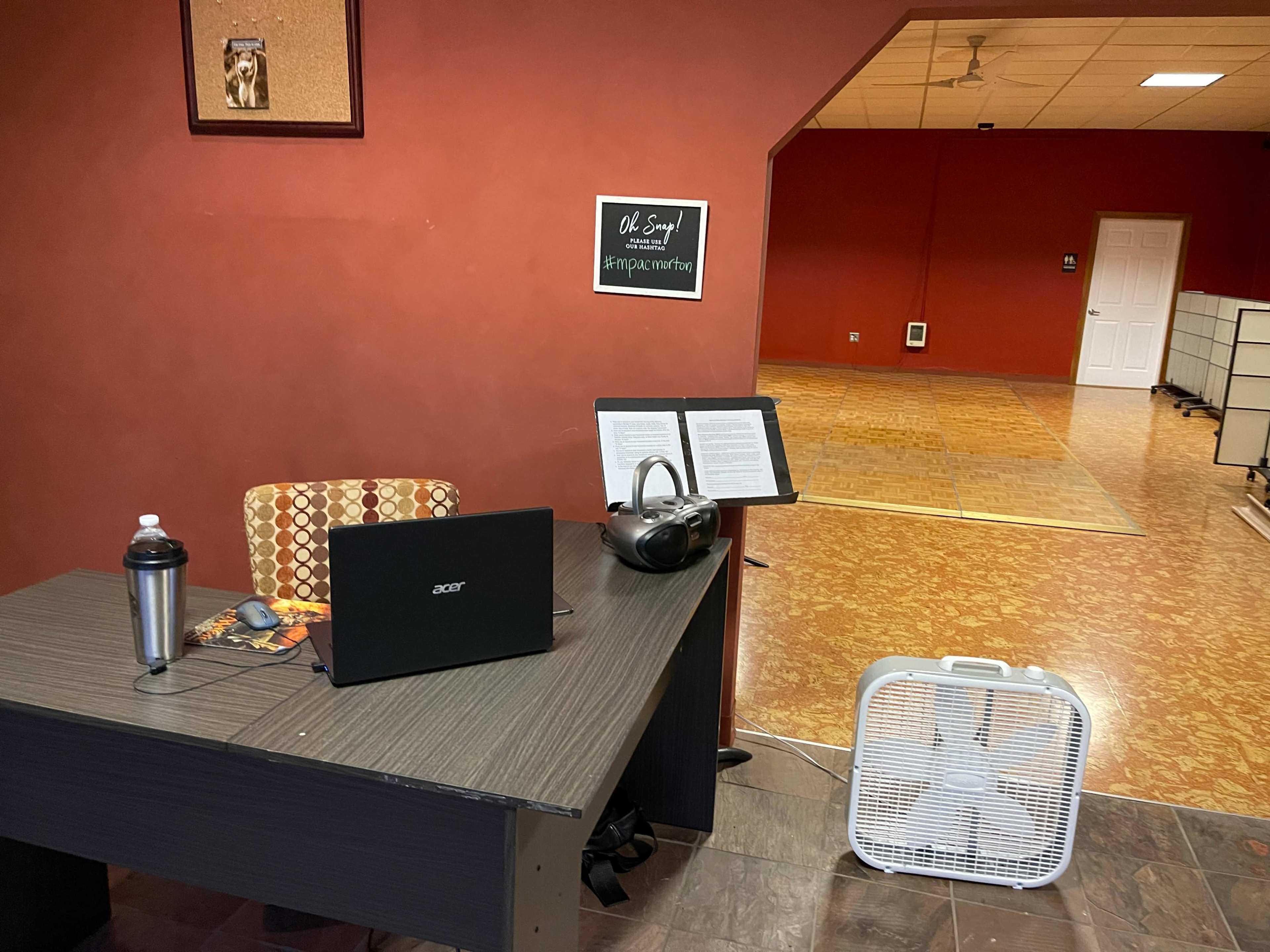 A computer desk with a laptop, headphones, and a fan is placed in a room with red walls and a large open space in the background.