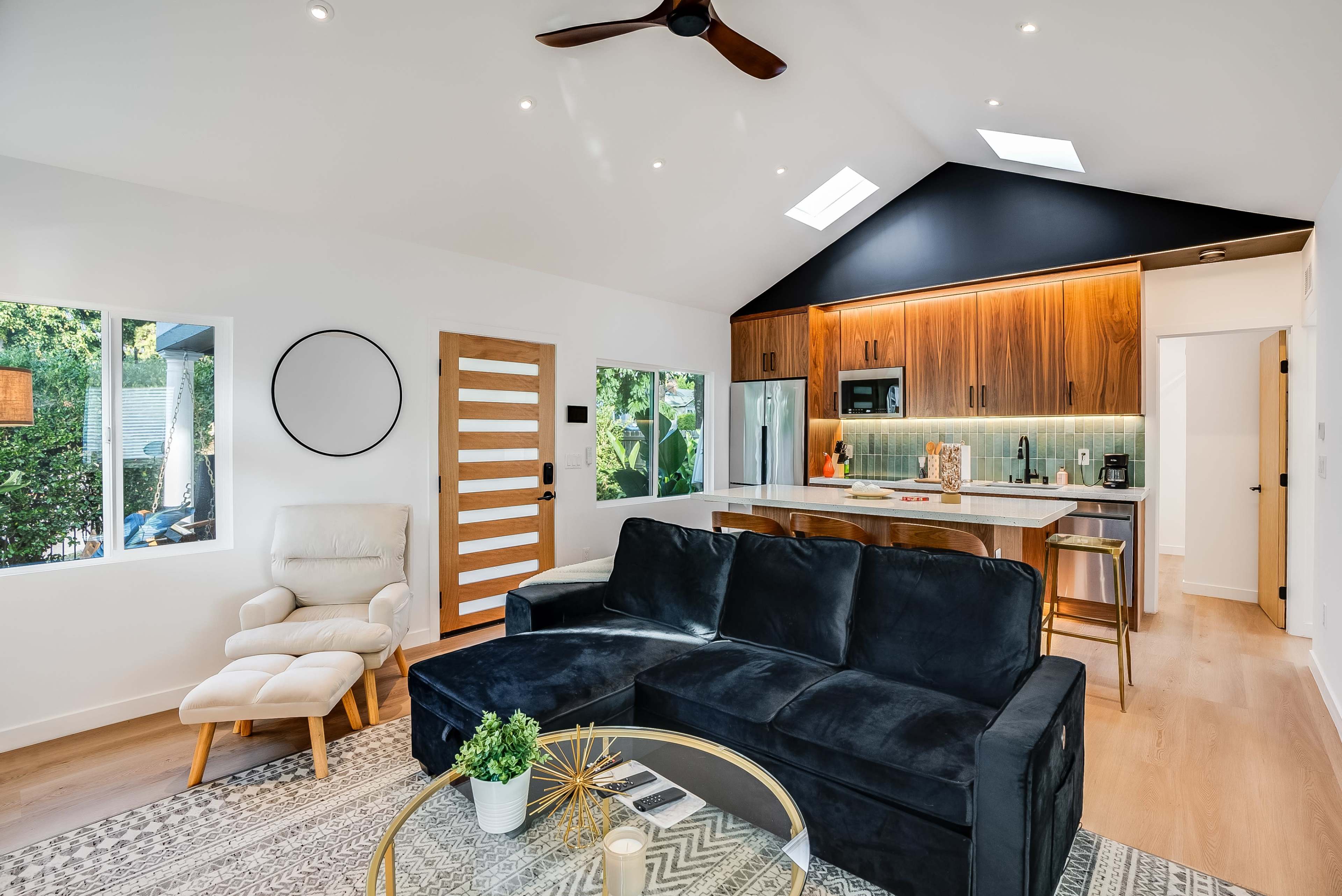 The image shows a modern living room with a black sectional sofa, a glass coffee table, a white armchair, and a kitchen visible in the background featuring wooden cabinets and bright lighting.