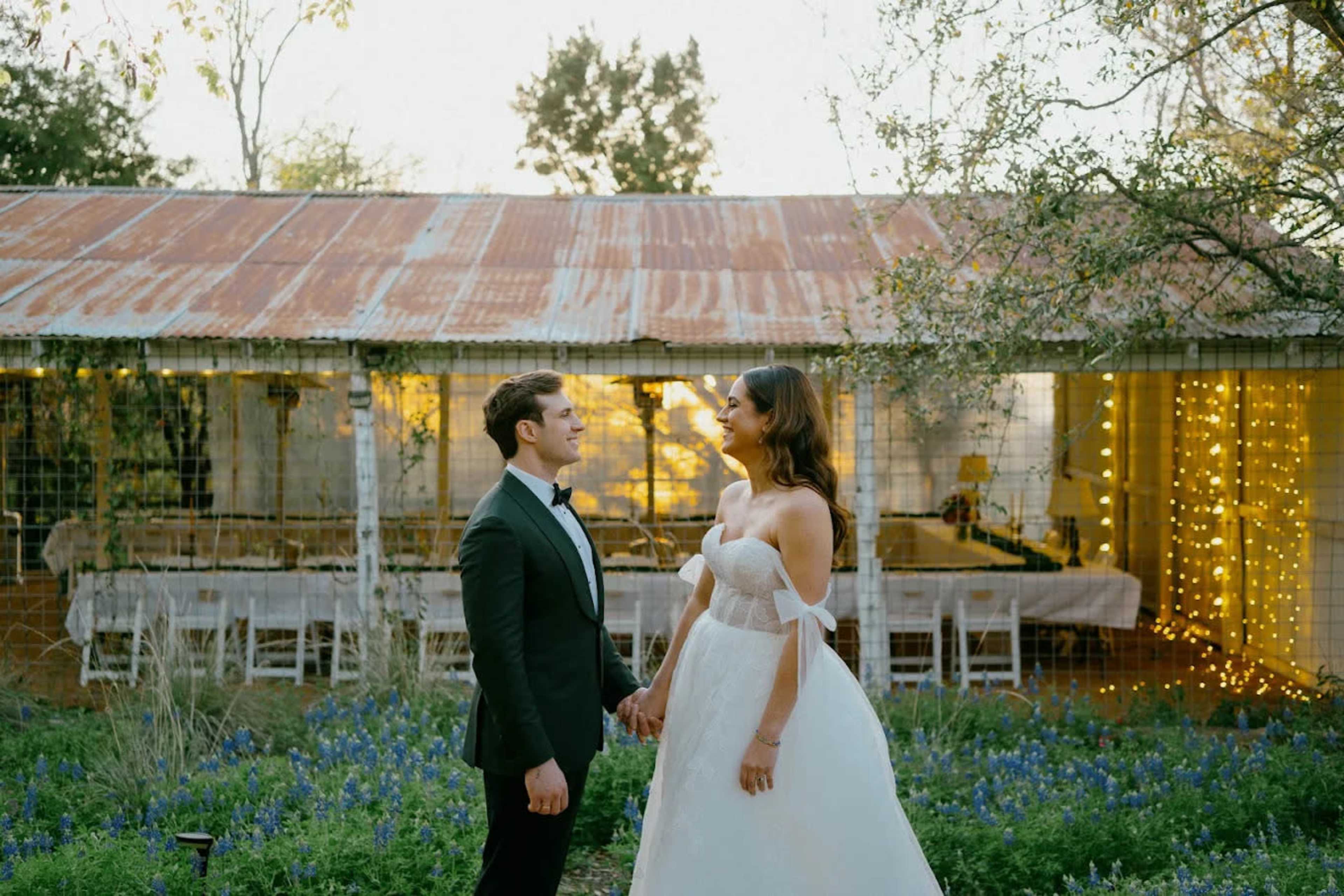A couple in formal attire stands hand in hand outside a rustic venue decorated with string lights and surrounded by flowers.