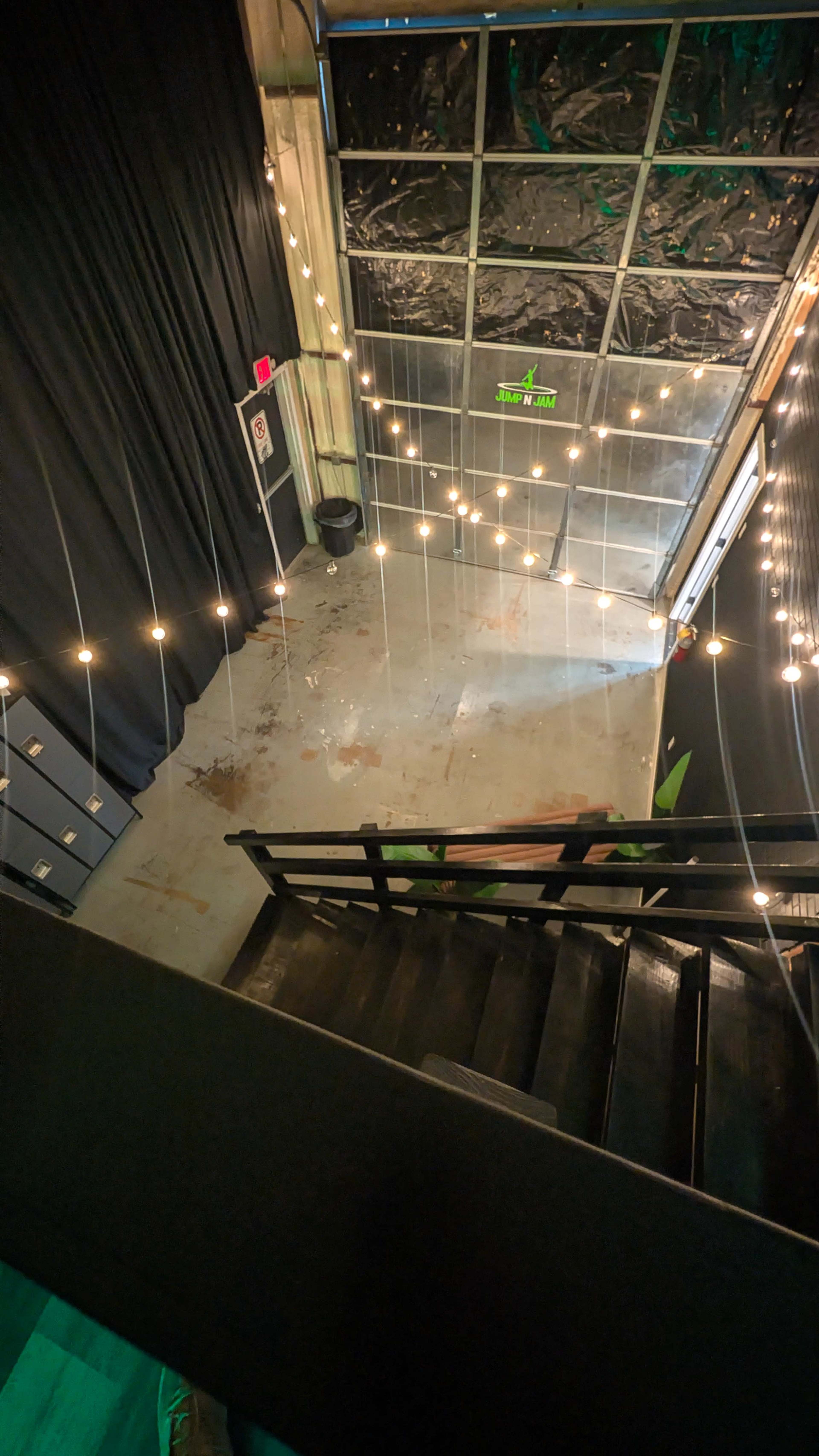 The image shows a stairway leading down to a dimly lit space with a concrete floor, adorned with string lights and a black curtain in the background.