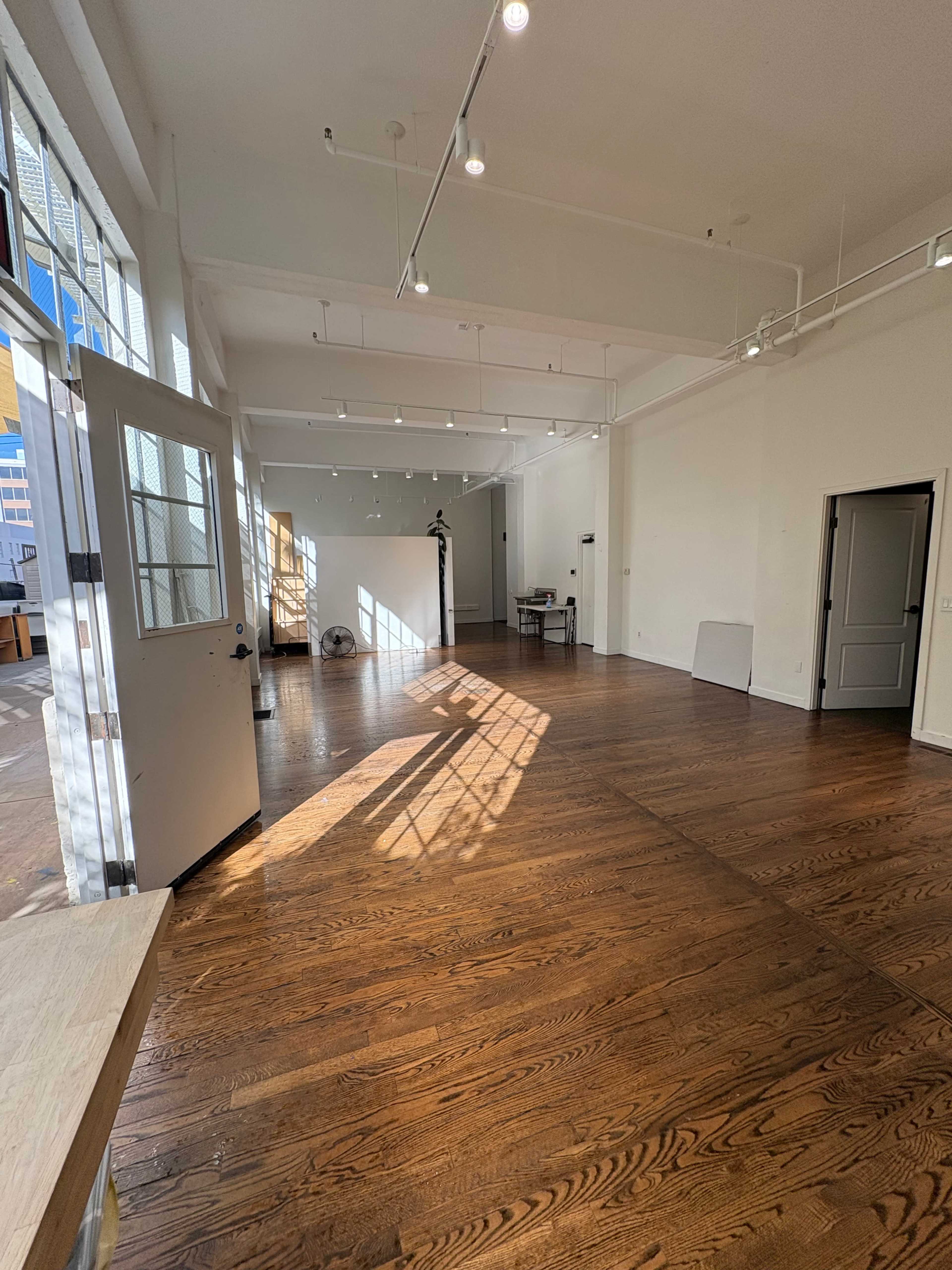 The image shows a spacious, well-lit interior with wooden floors, large windows, and minimalist decor.