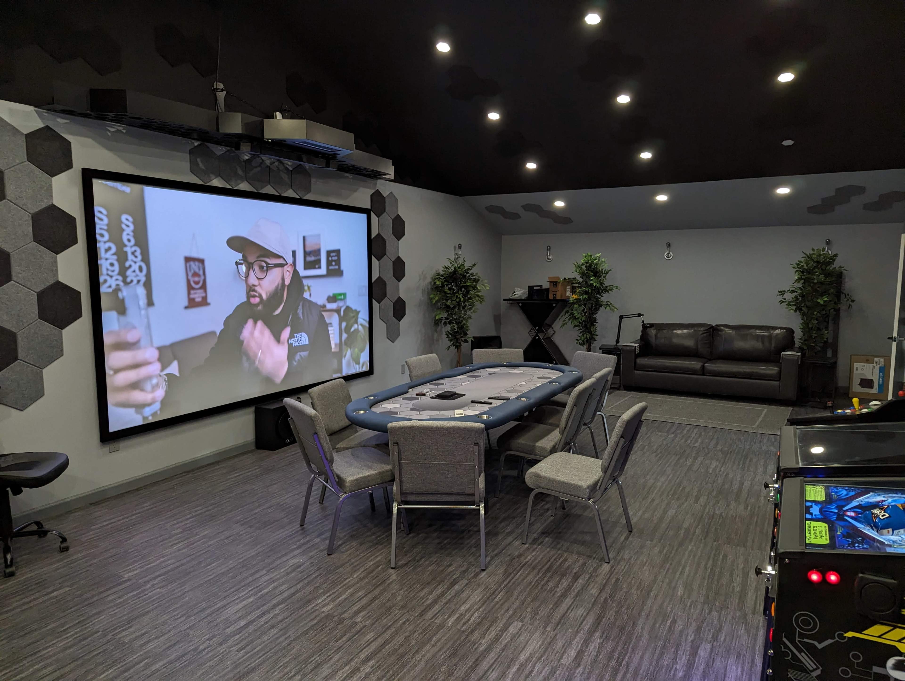 A game room with a large projection screen displaying a person, a poker table surrounded by chairs, and a seating area with a couch.